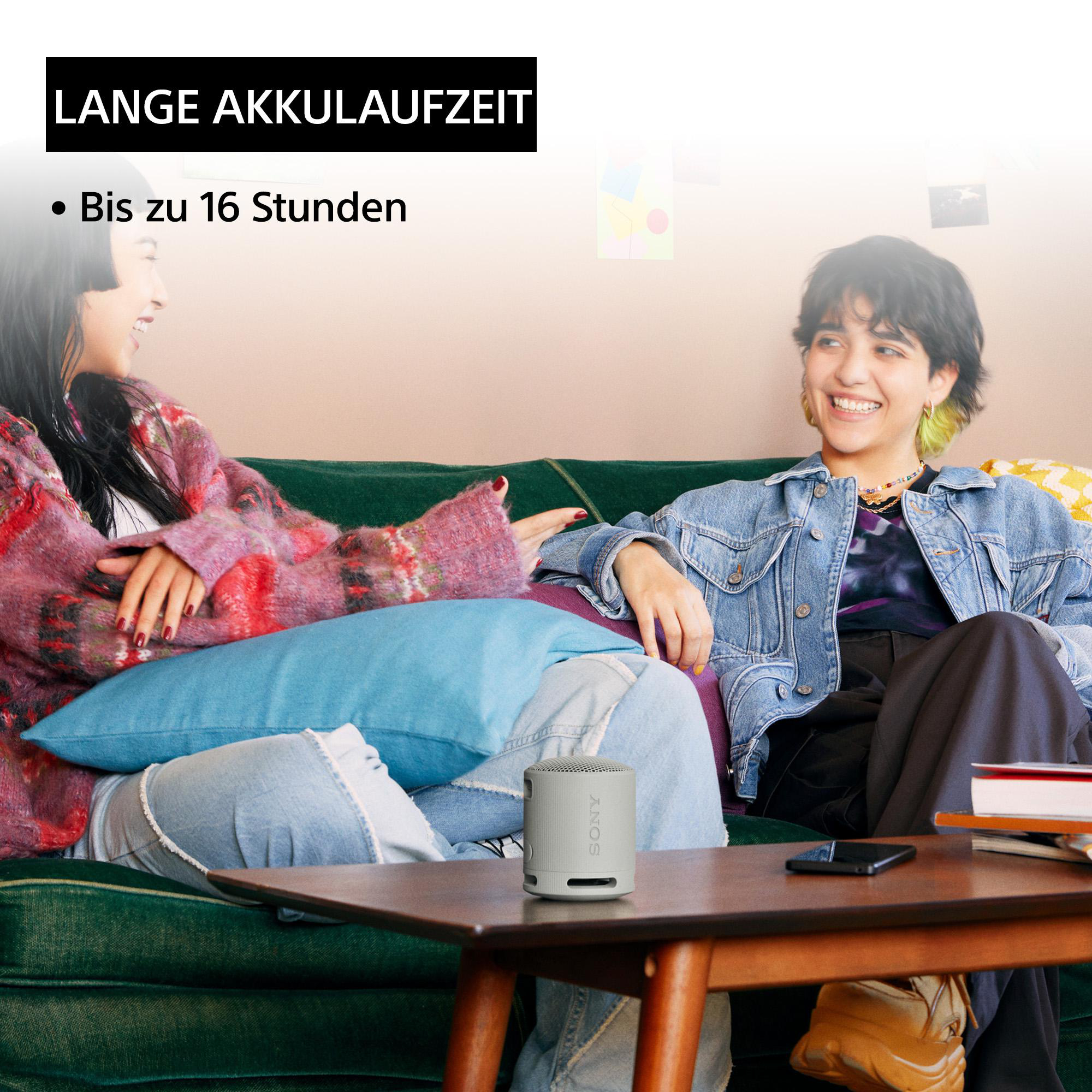 Zwei Frauen sitzen auf einer grünen Couch, lächeln, ein kleiner grauer Lautsprecher.