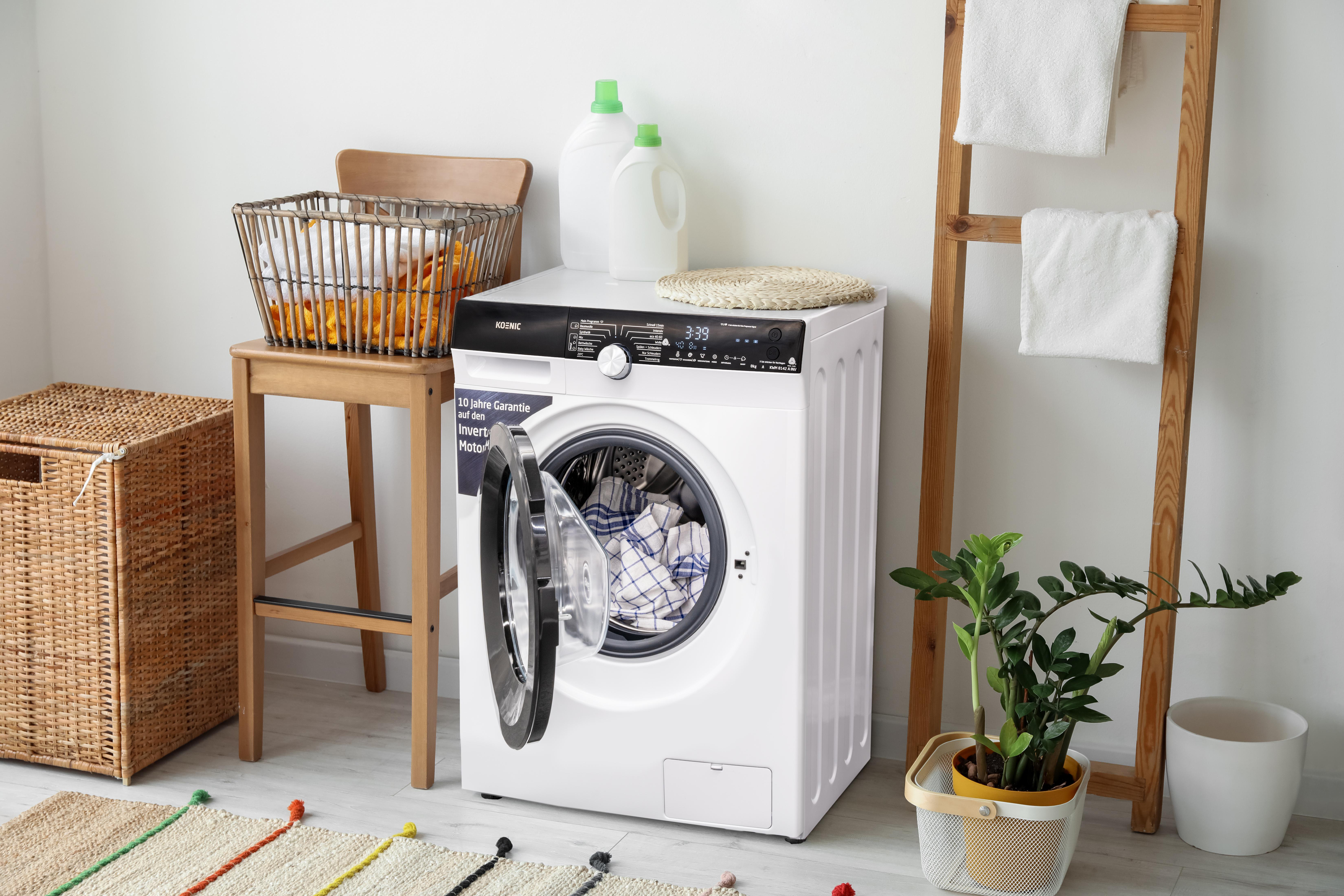 Weiße Waschmaschine mit offener Tür. Ein Korb, ein Hocker, Pflanzen und Handtücher auf einer Holzleiter sind sichtbar.