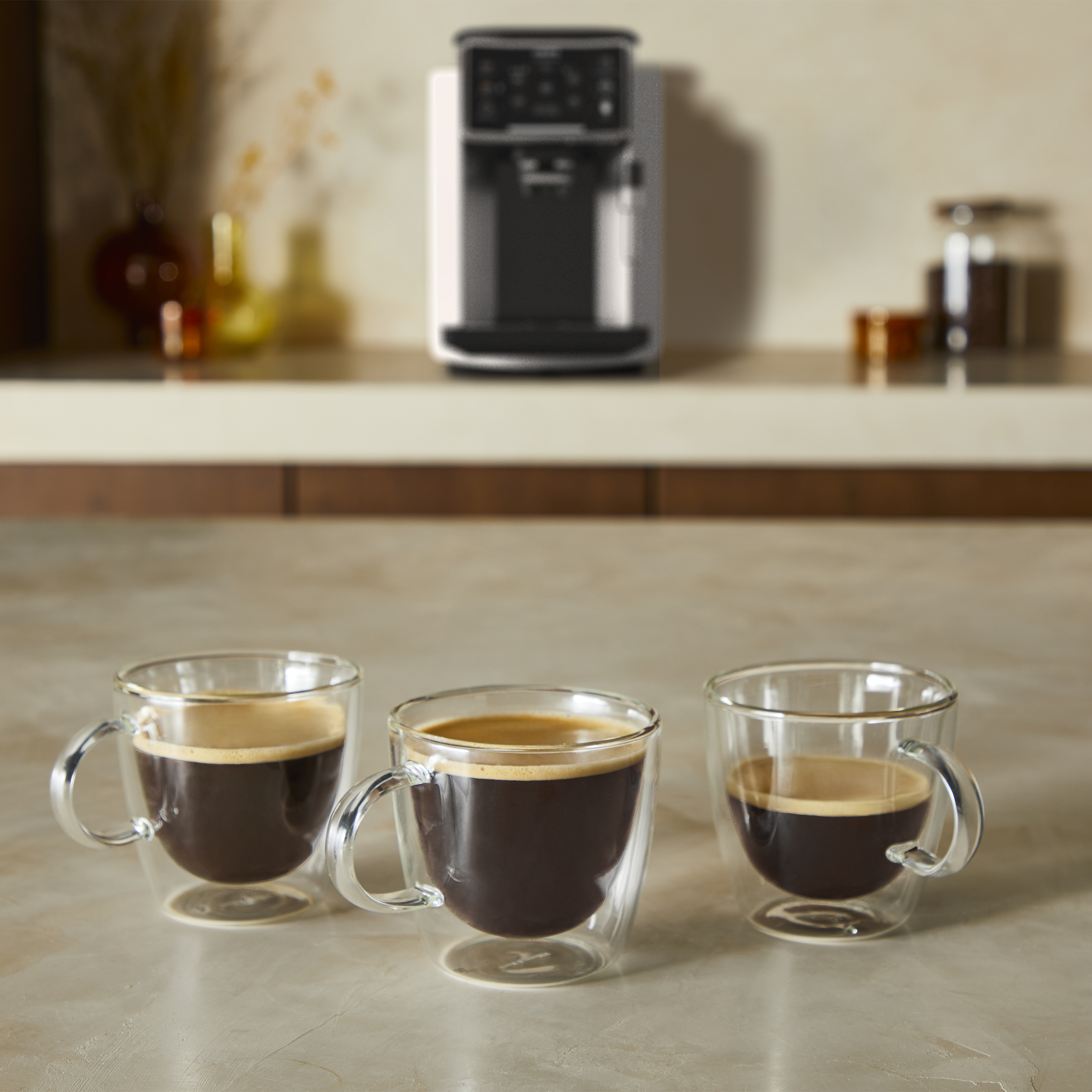 Drei Kaffeetassen mit dunklem Kaffee auf einer hellen Arbeitsplatte in der Nähe einer Kaffeemaschine.