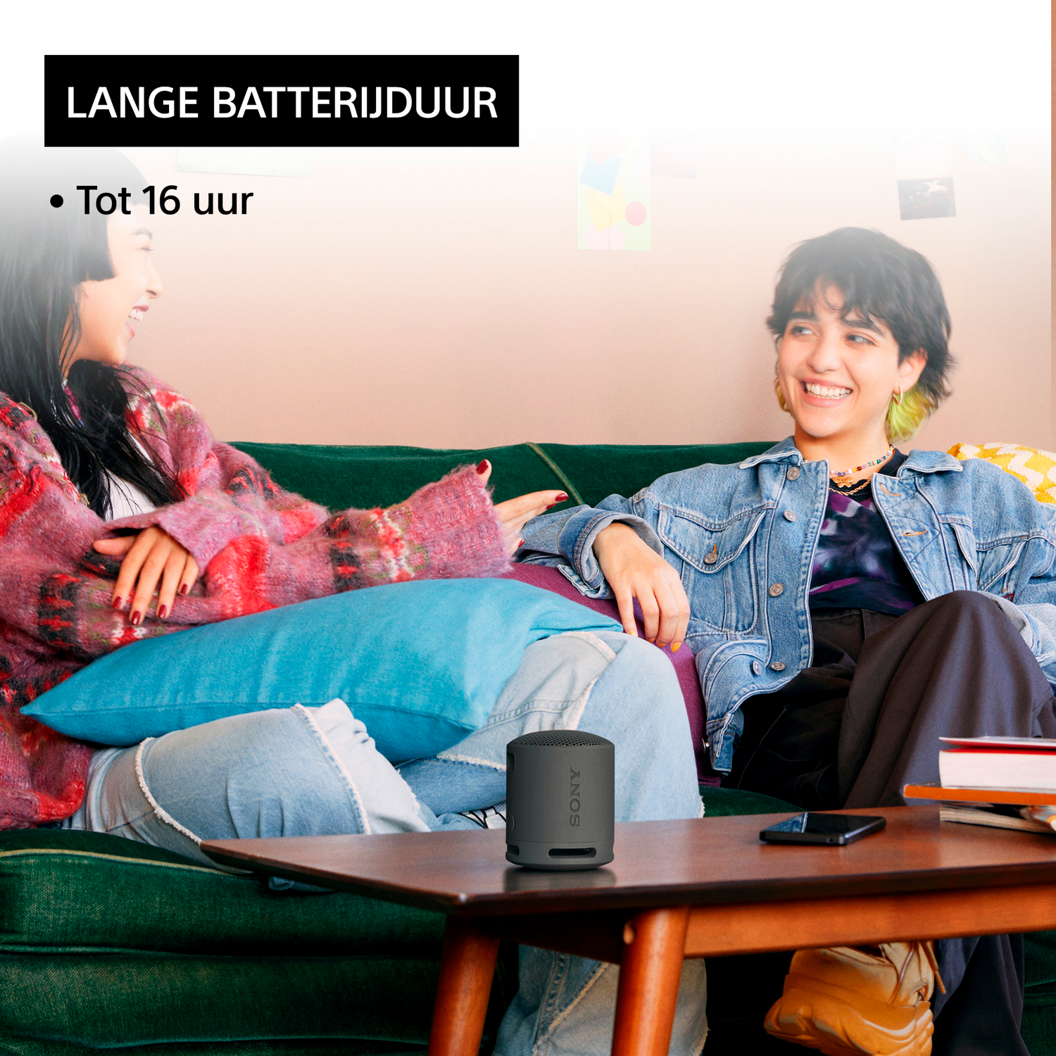 Twee vrouwen lachen, zitten op een groene bank. Een zwarte luidspreker staat op een tafel. Tekst: Lange batterijduur.
