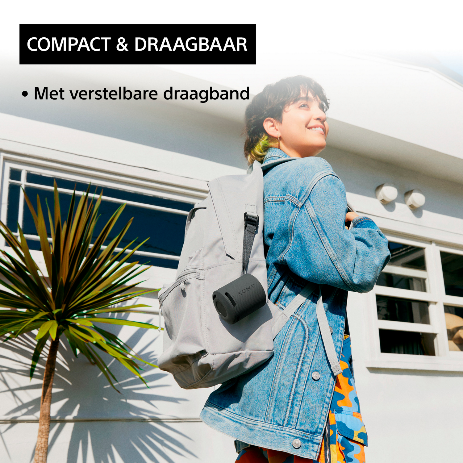 Vrouw met rugzak en speaker, buiten wit gebouw met palmboom.