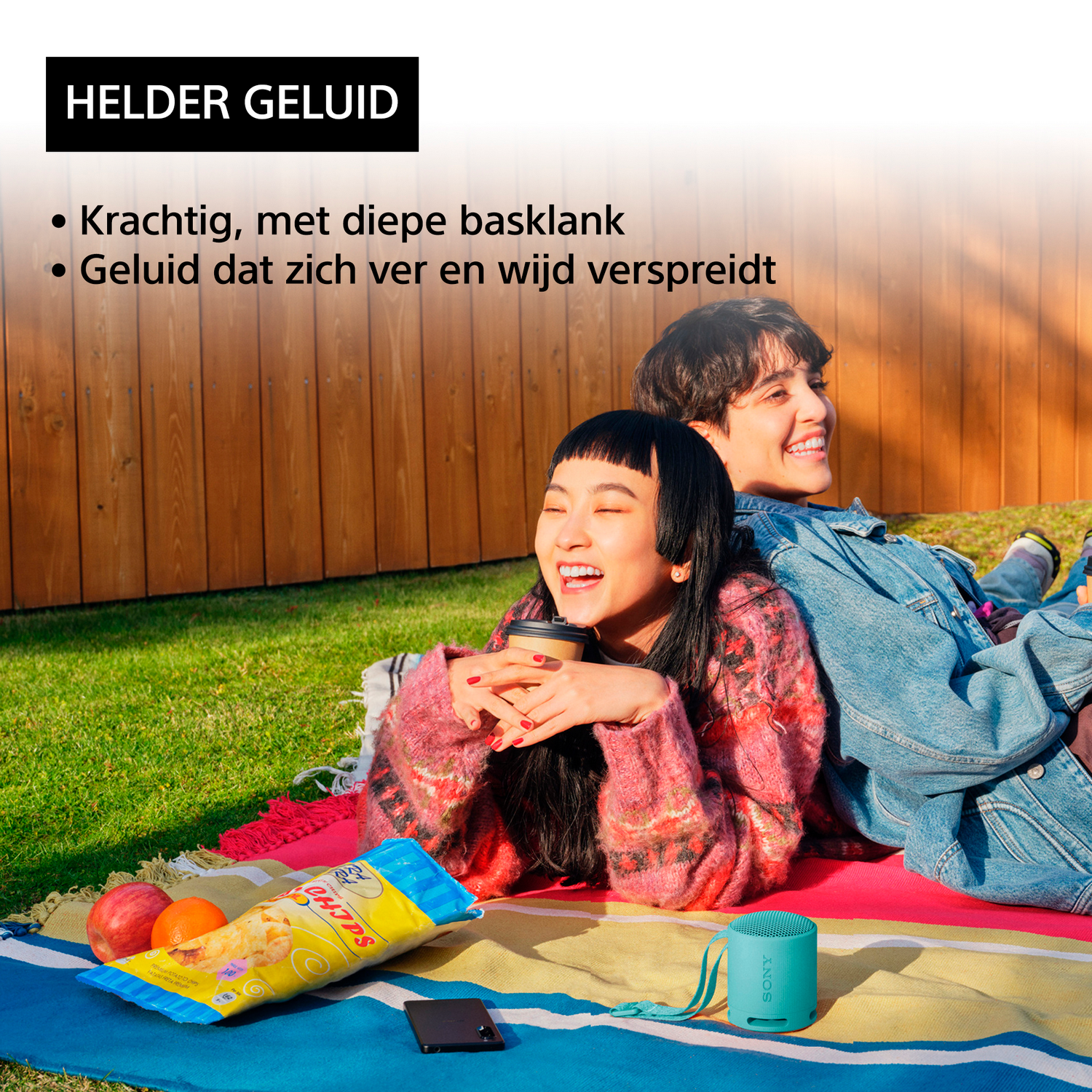 Two people sitting on a blanket; one is holding a drink and the other is smiling.