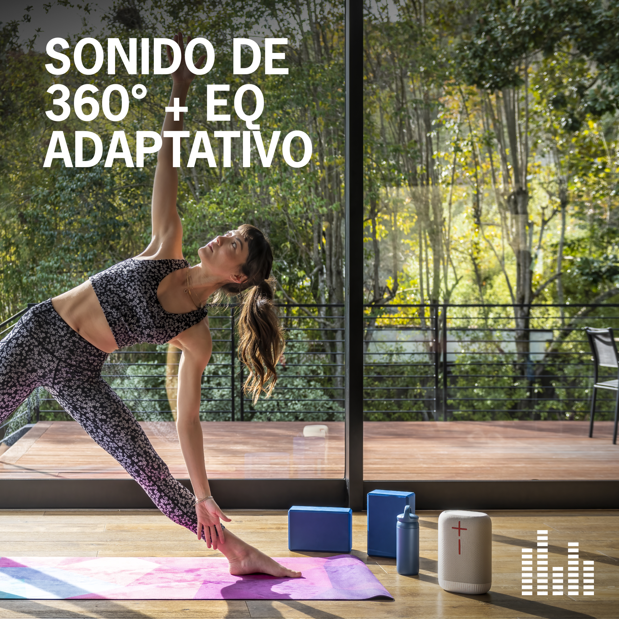 Mujer en pose de yoga, con altavoz, bloques y esterilla. Fondo con pared de cristal y árboles.