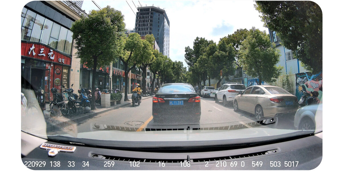 Widok z kamery samochodowej na ulicę z samochodami, motocyklami i budynkami.