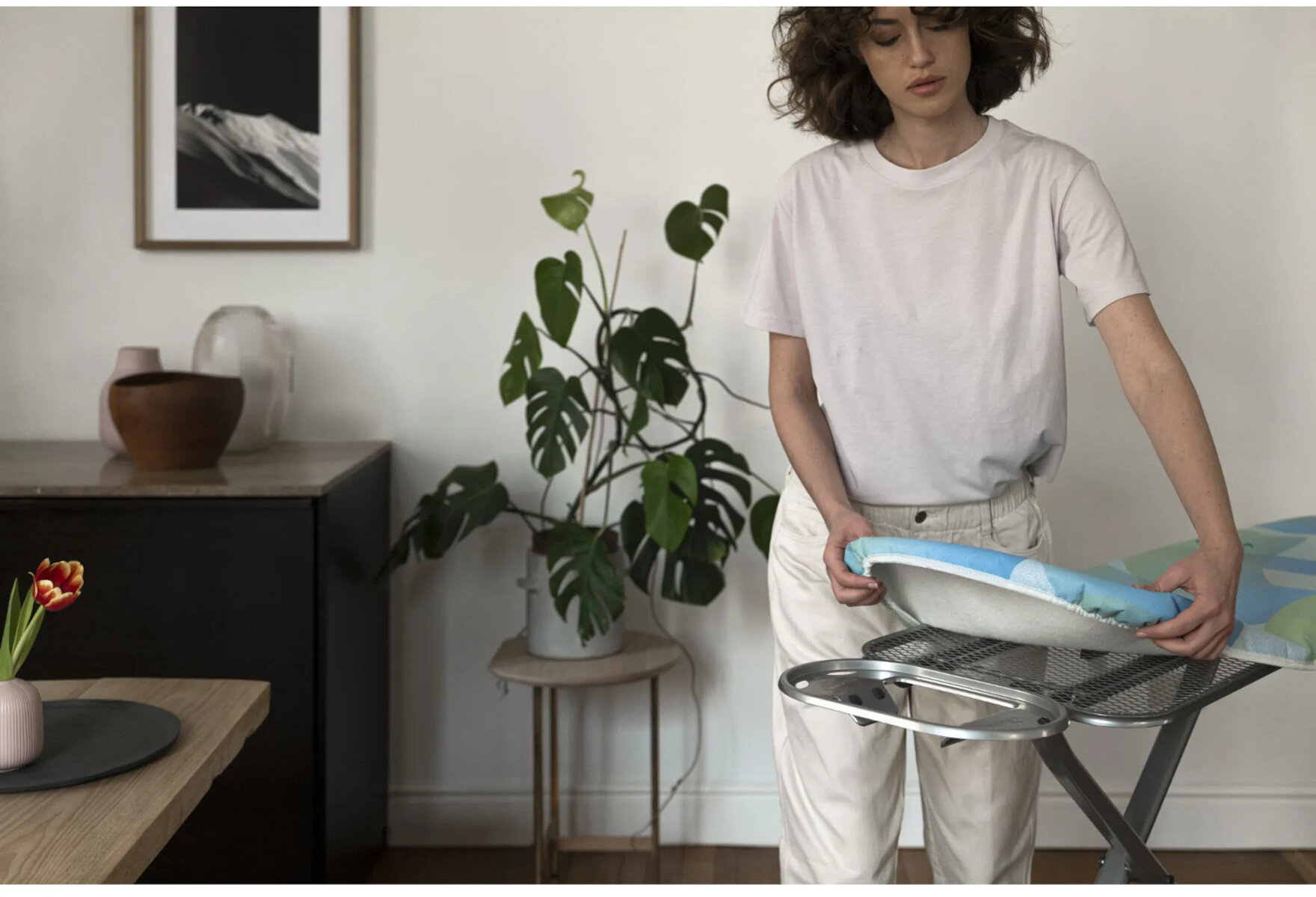 Kobieta rozkłada deskę do prasowania. Roślina doniczkowa i dzieła sztuki w tle pokoju.