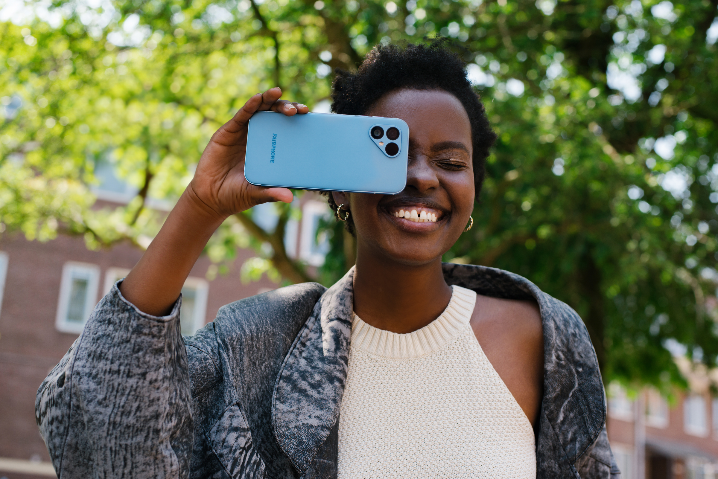 Eine Frau, die ein blaues Telefon über ihrem Auge hält, lächelt mit einem Stadtbild im Hintergrund.