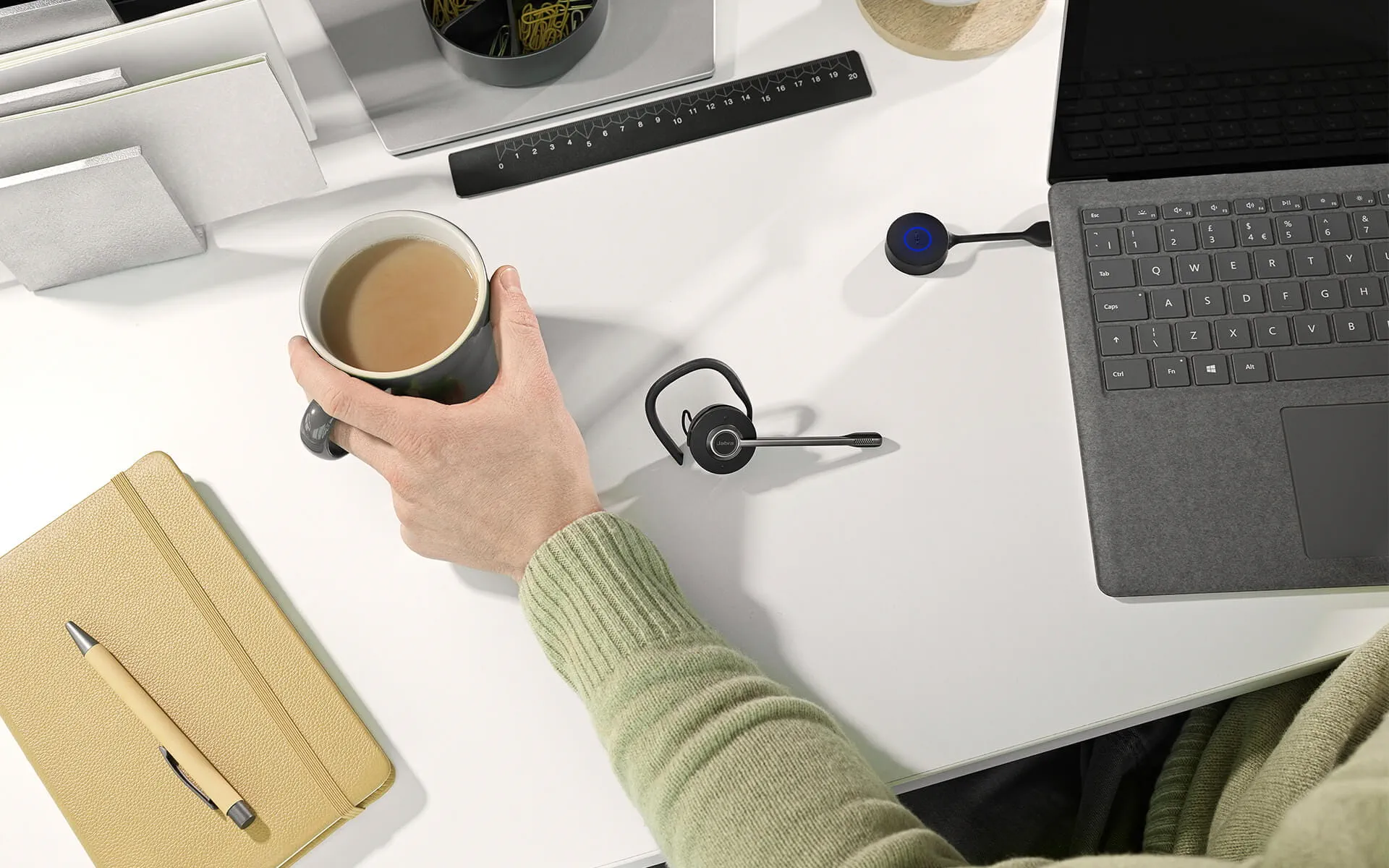 Eine Person hält eine Tasse mit Kaffee, Headset auf einem Tisch, Laptop offen, Stift und Notizbuch.
