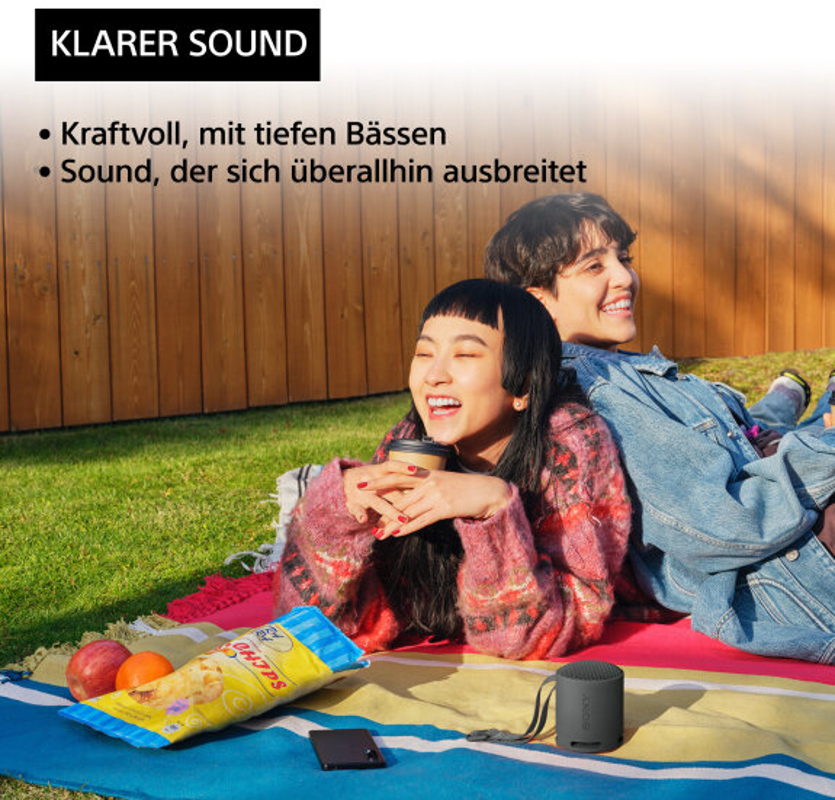Zwei Personen sitzen lachend auf einer Wiese. Ein kleiner schwarzer Lautsprecher, Snacks und ein Telefon liegen daneben.