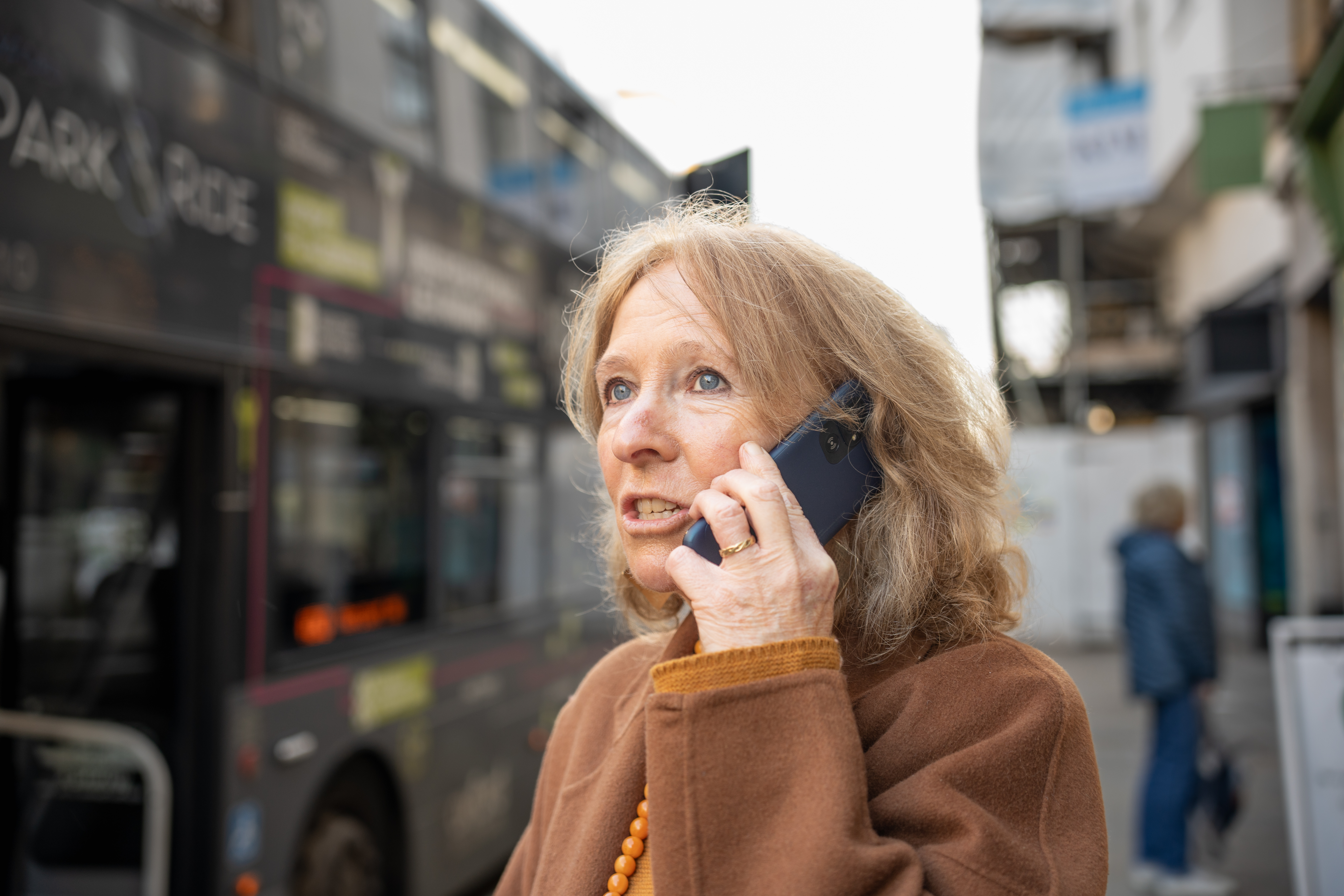 Une femme âgée au téléphone se tient près d'un bus. Elle a l'air préoccupée.