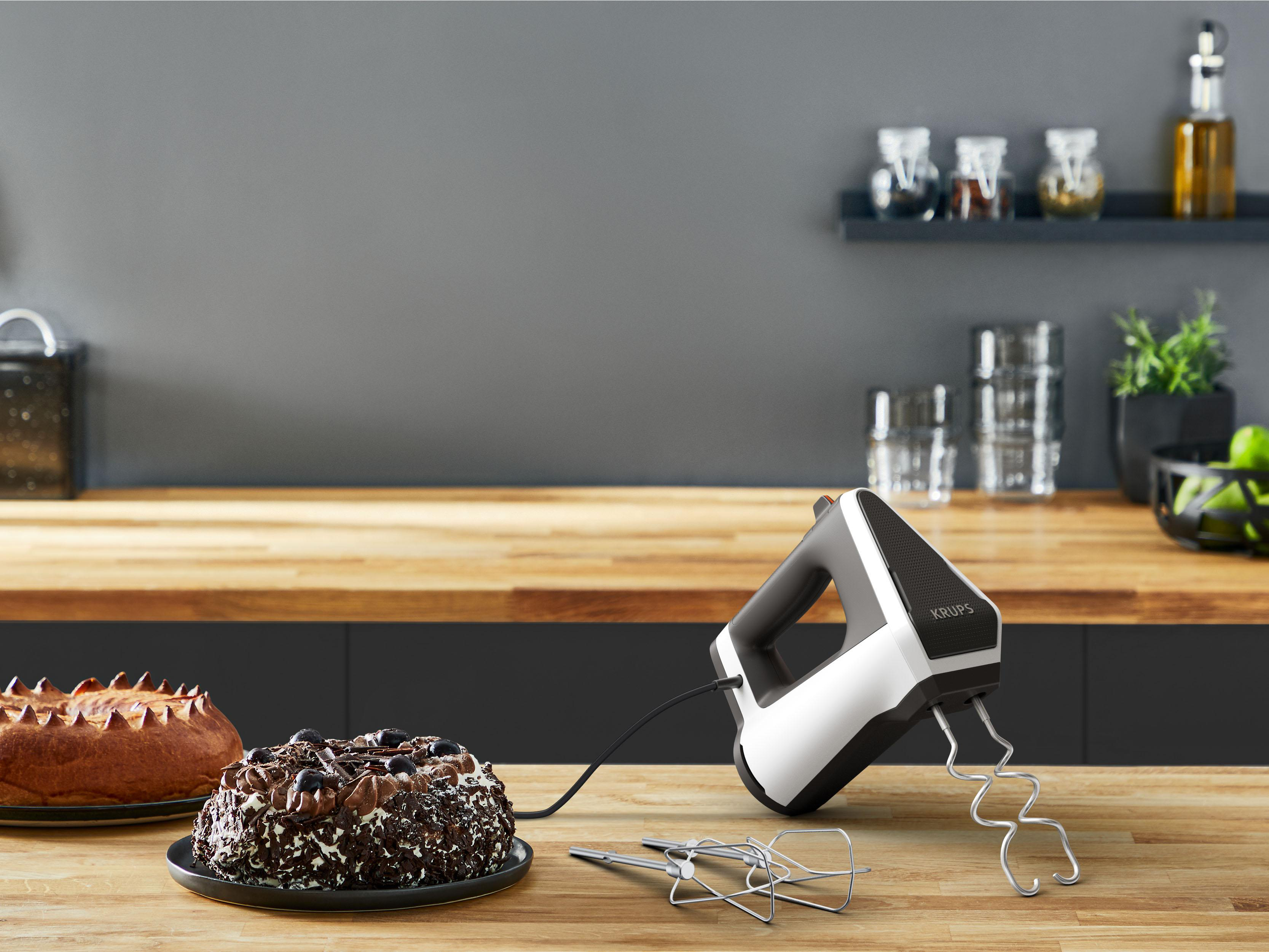 Ein Handmixer auf einer Holztheke mit zwei Kuchen, einer schokoladenüberzogen, und anderem Zubehör im Hintergrund.