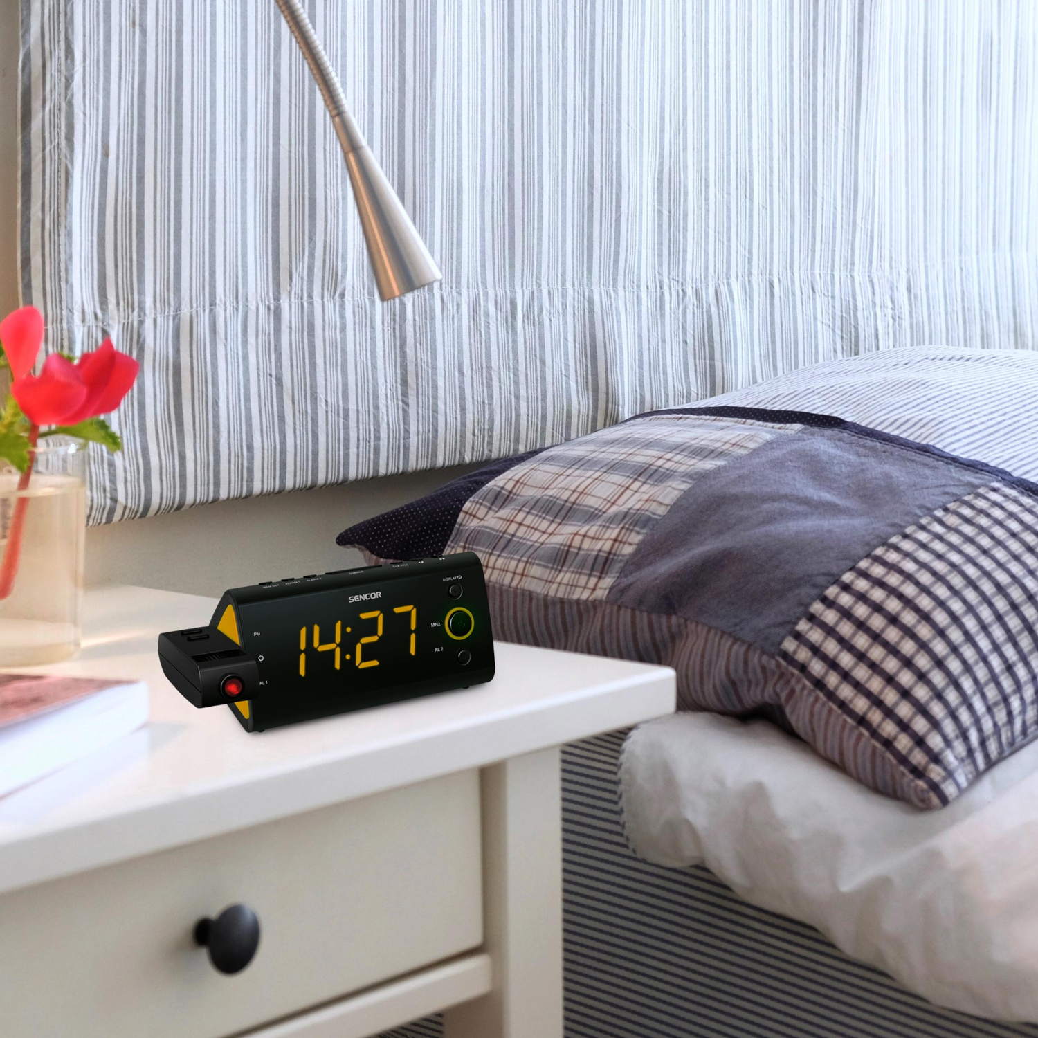 Fekete digitális óra 14:27-et mutat egy fehér asztalon, virággal és ággyal.