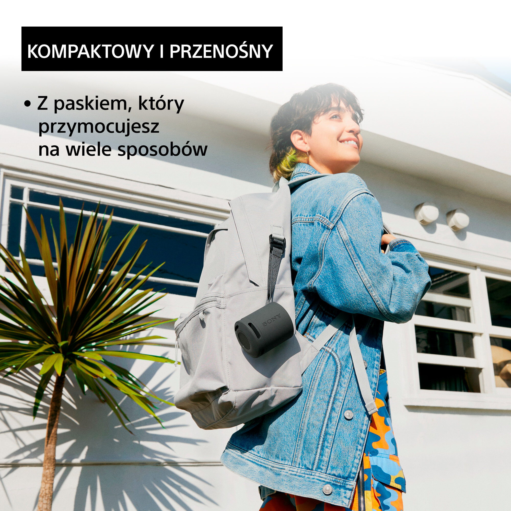 Kobieta z plecakiem i czarnym głośnikiem, obok palmy.