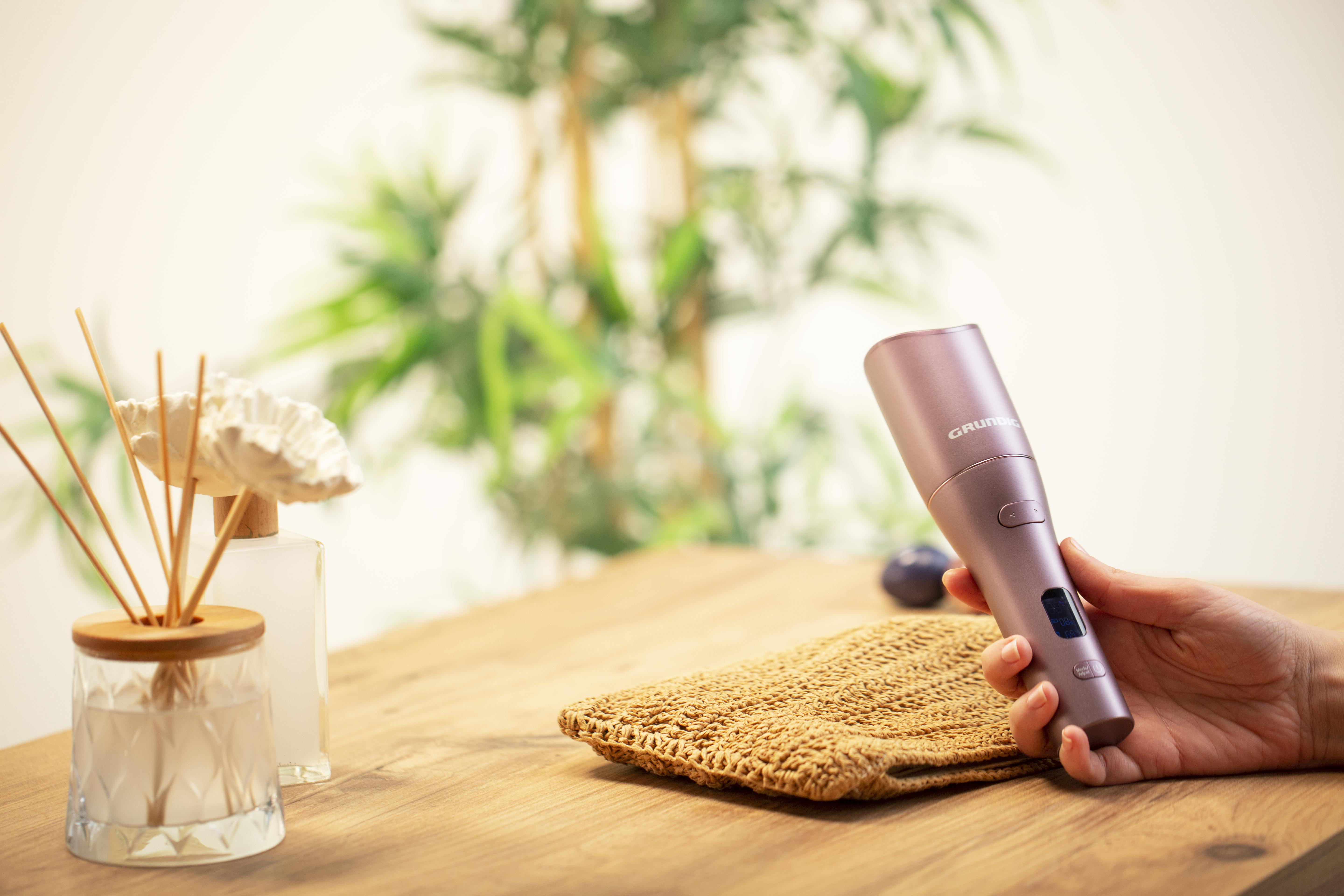 Eine Person hält ein rosa Grundig-Gerät auf einem Holztisch. Dekorative Objekte befinden sich im Hintergrund.