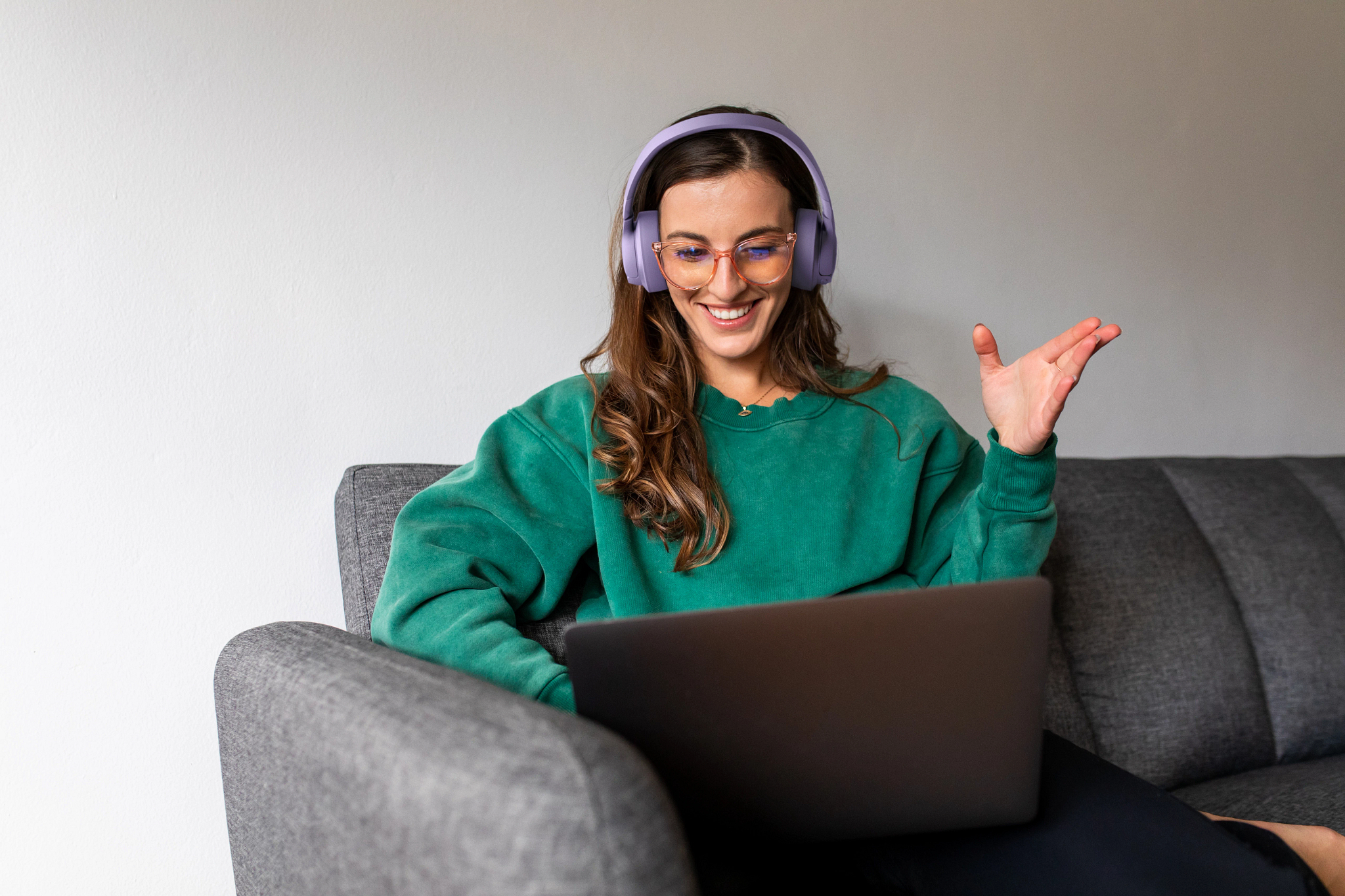 Eine Frau mit Kopfhörern und Laptop lächelt und gestikuliert auf einem grauen Sofa.