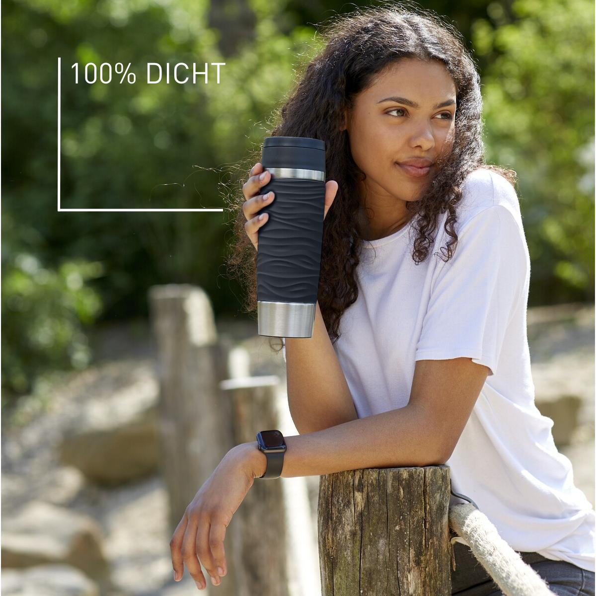 Frau hält einen schwarzen Thermobecher, Text lautet „100 % Dicht“, in einer Umgebung im Freien.