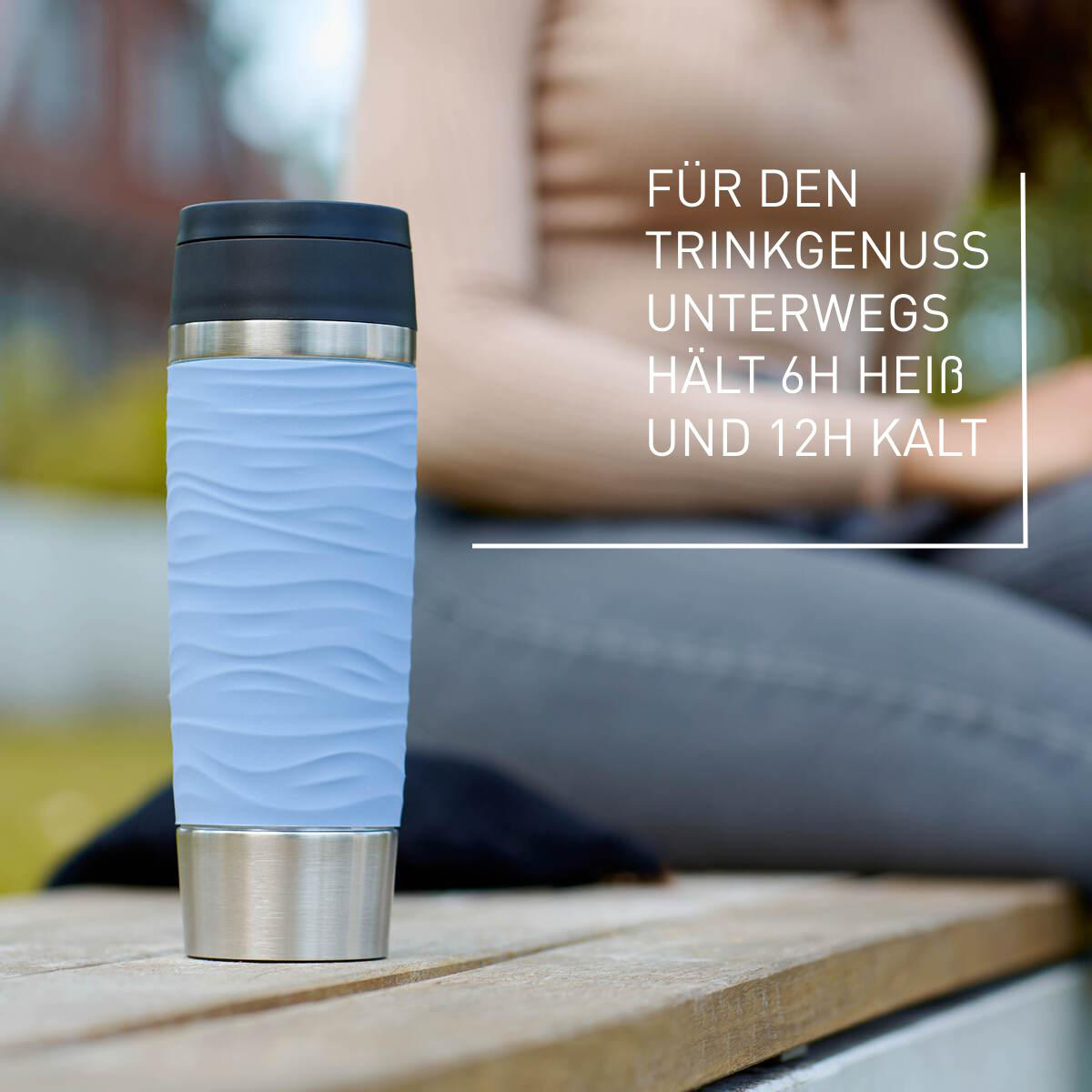 Ein hellblauer Thermobecher mit Wellenlinien steht auf einer Holzoberfläche. Text sagt, er hält warm oder kalt.