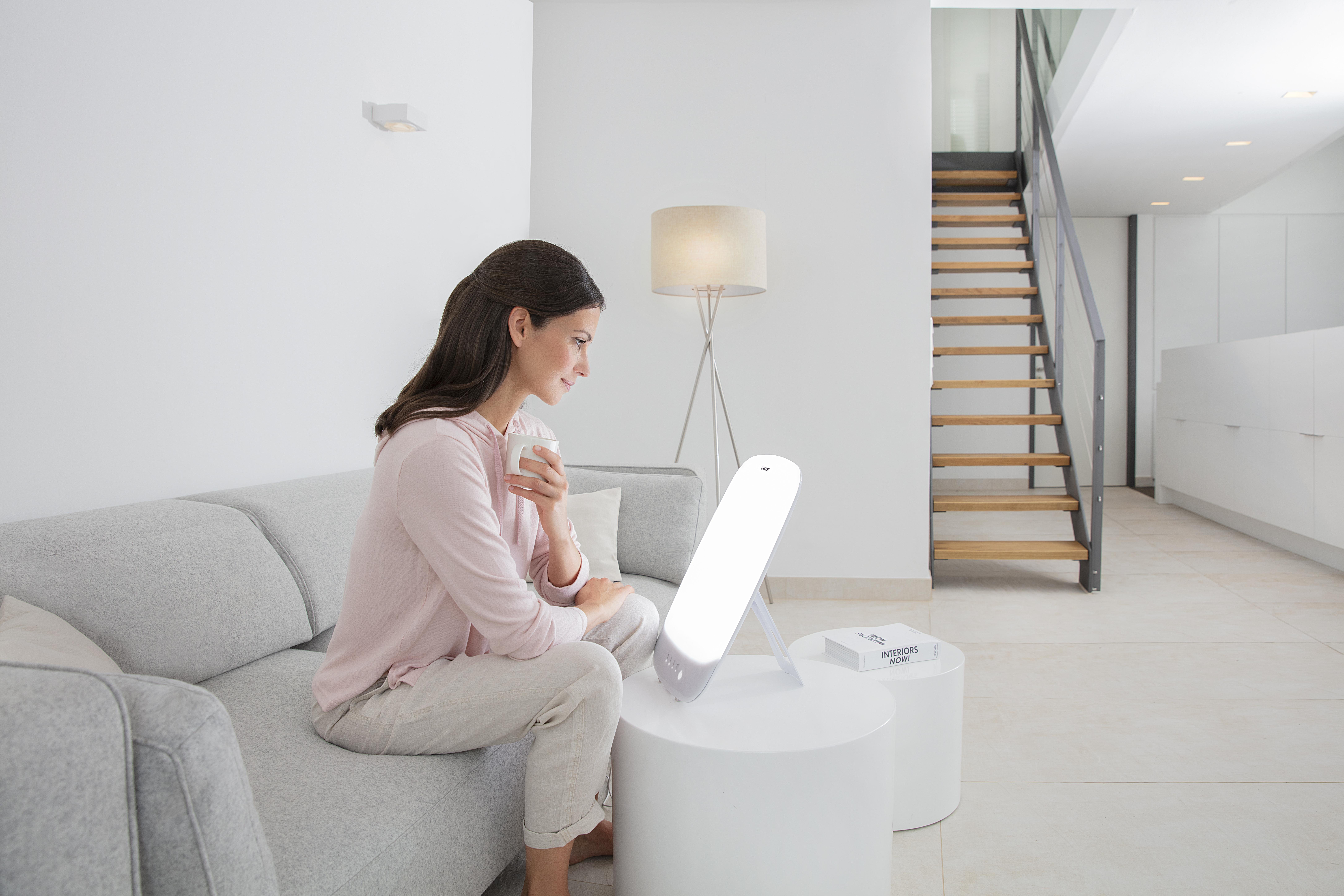 Eine Frau sitzt auf einem grauen Sofa, hält eine Tasse und schaut auf eine Lichttherapielampe.