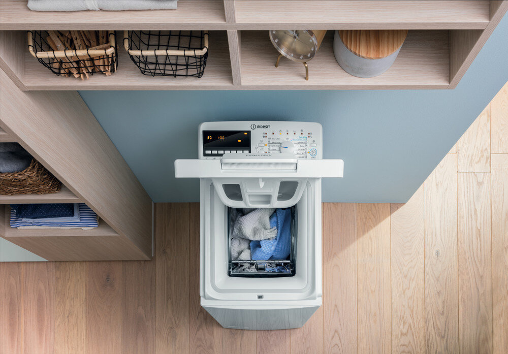 Widok z góry na pralkę, z otwartą pokrywą, praniem w środku i półkami w tle.