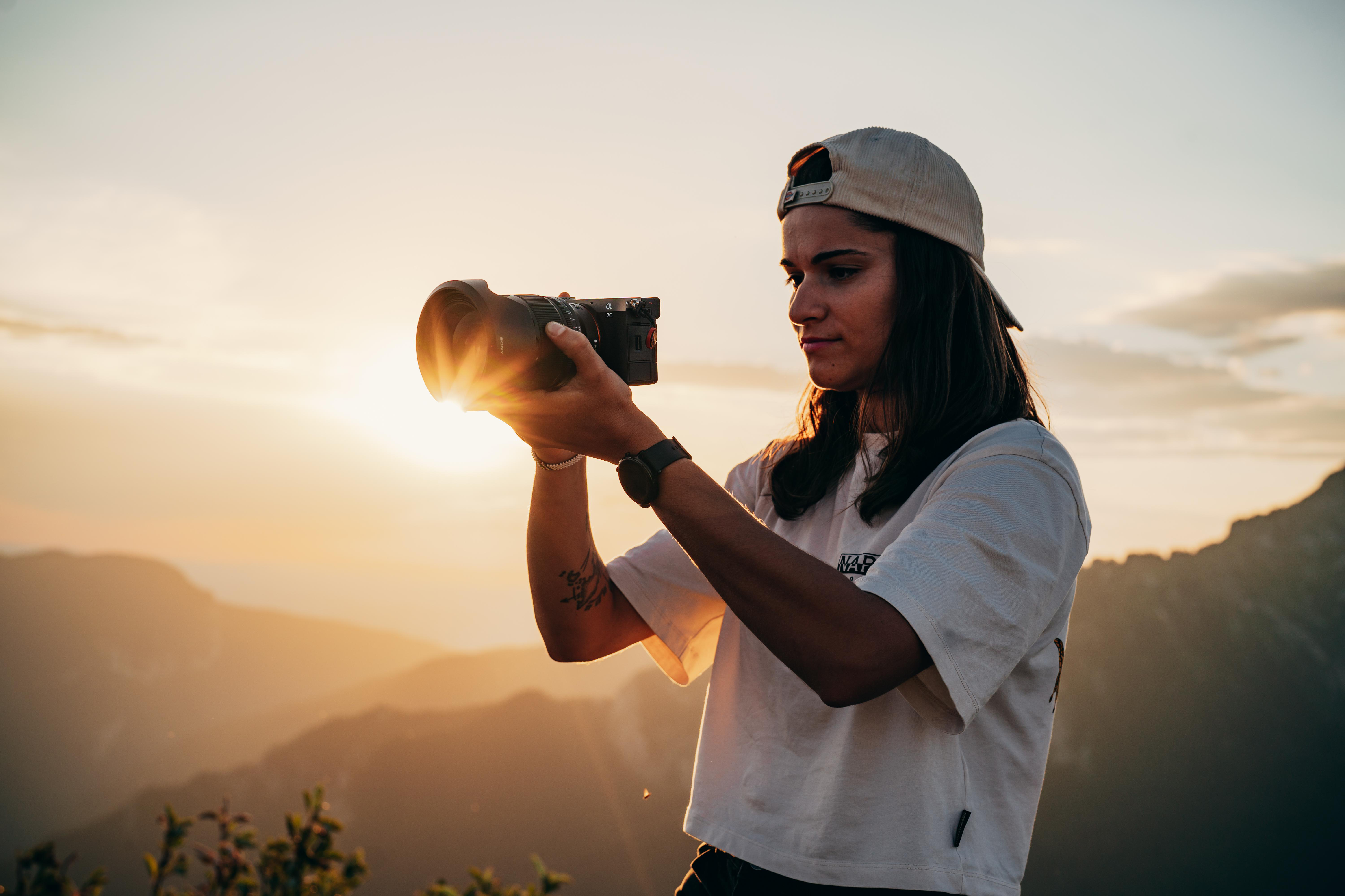 Frau hält eine Kamera und macht Fotos bei Sonnenuntergang, mit Bergen im Hintergrund.