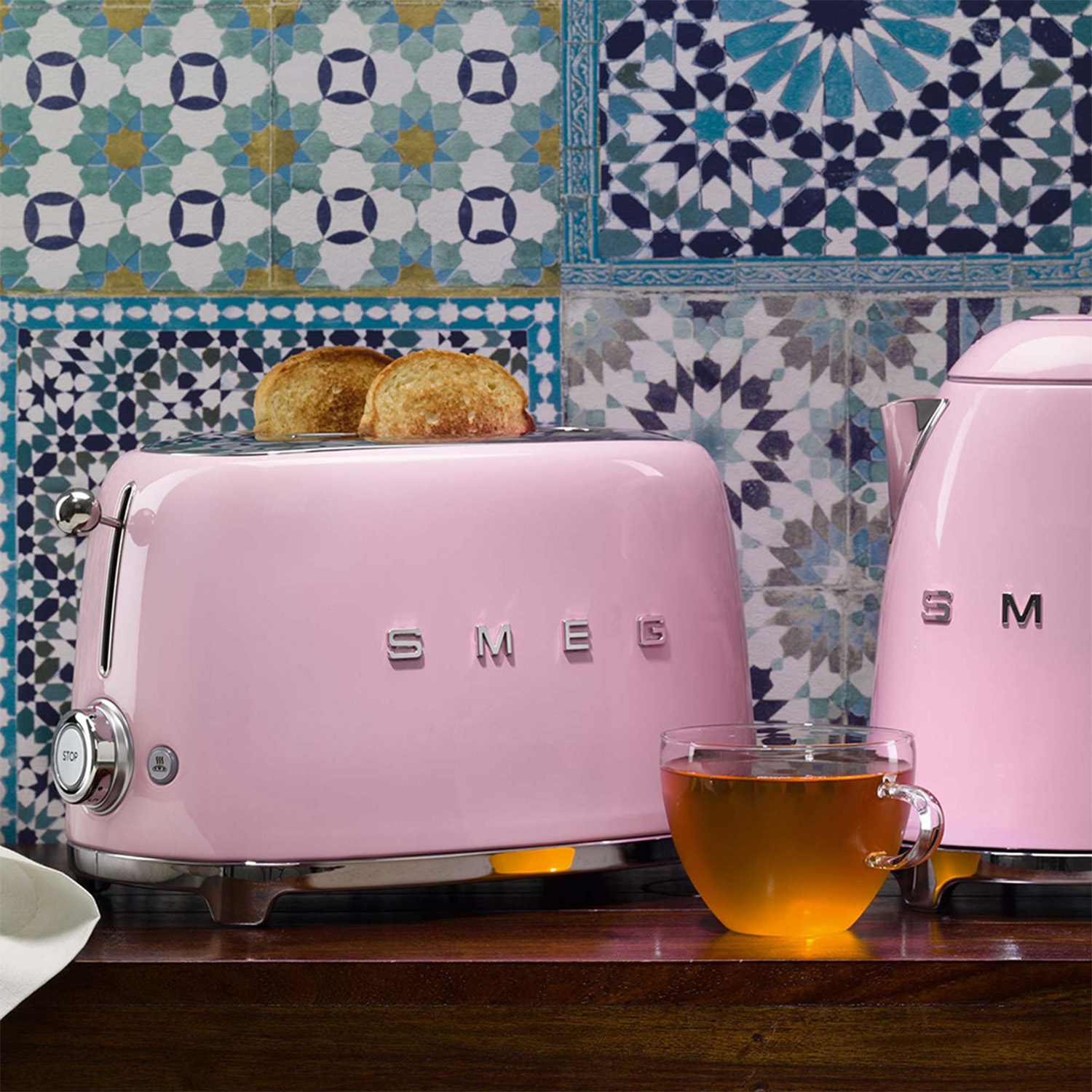 Un tostapane SMEG rosa con pane tostato, un bollitore rosa e un bicchiere di tè su una superficie di legno.