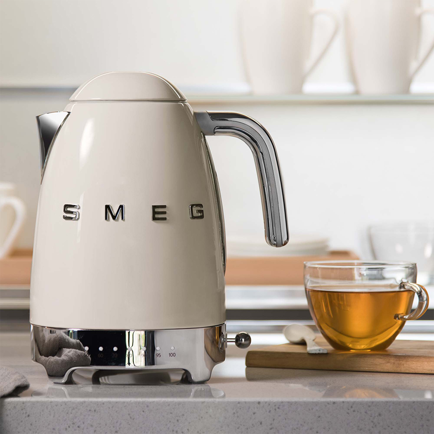 Un bollitore elettrico Smeg color crema con manico e base cromati, accanto a un bicchiere di tè e un tagliere.