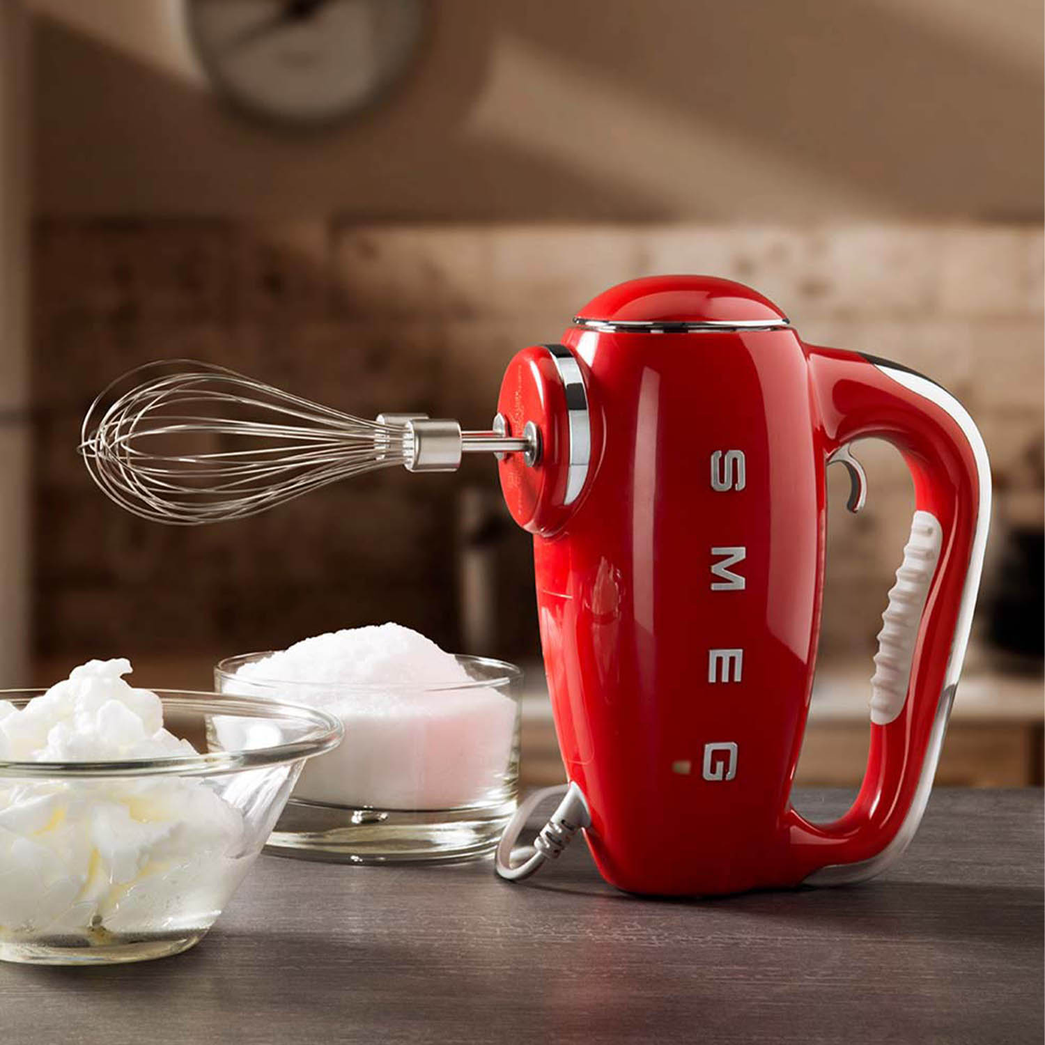Frullatore a mano SMEG rosso con frusta, accanto a ciotole di zucchero e panna montata su una superficie di legno.