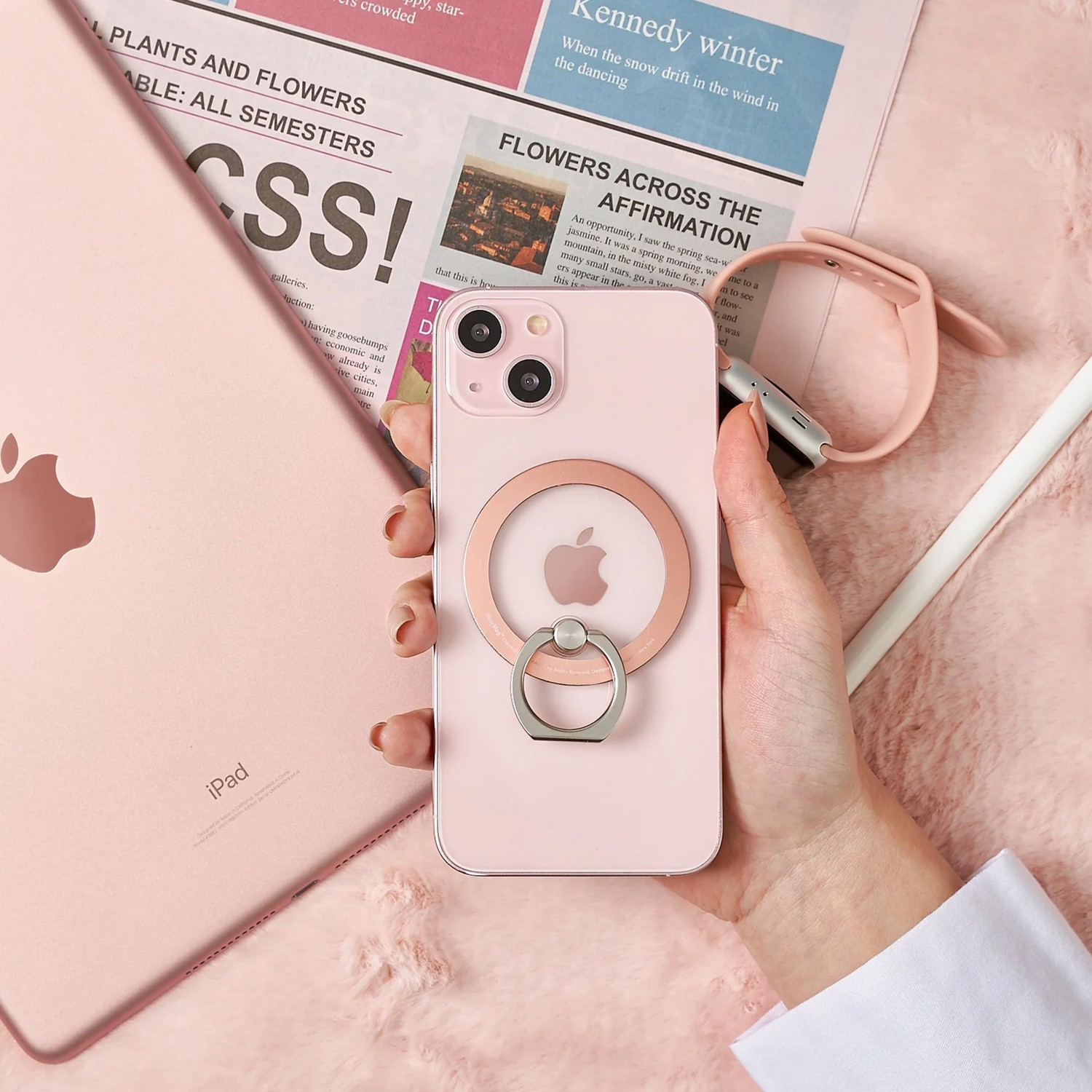Téléphone rose avec support de bague tenu par une main, à côté d'un iPad, sur une surface rose avec une montre.