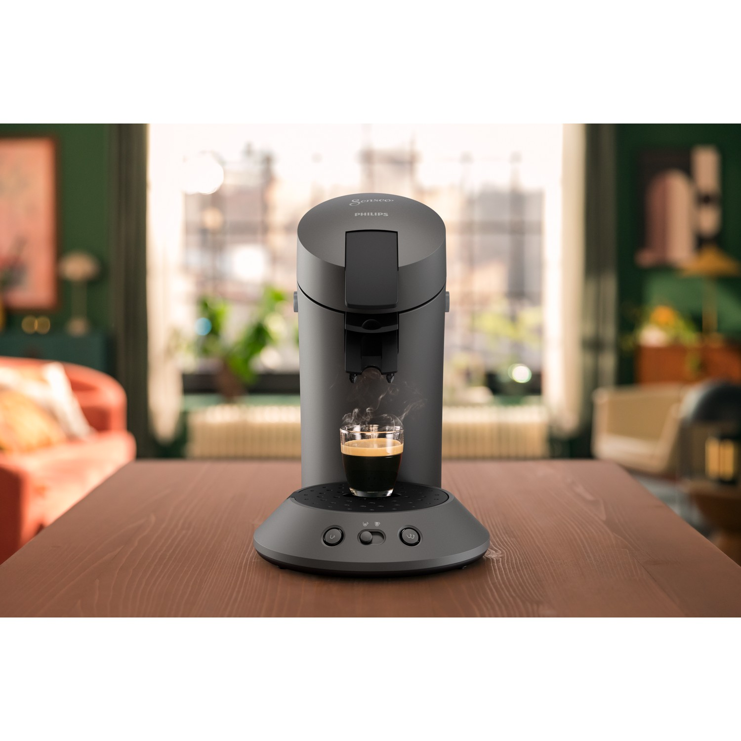 A dark gray Philips Senseo coffee machine with a cup of espresso on a wooden table. Background shows a room with a window.