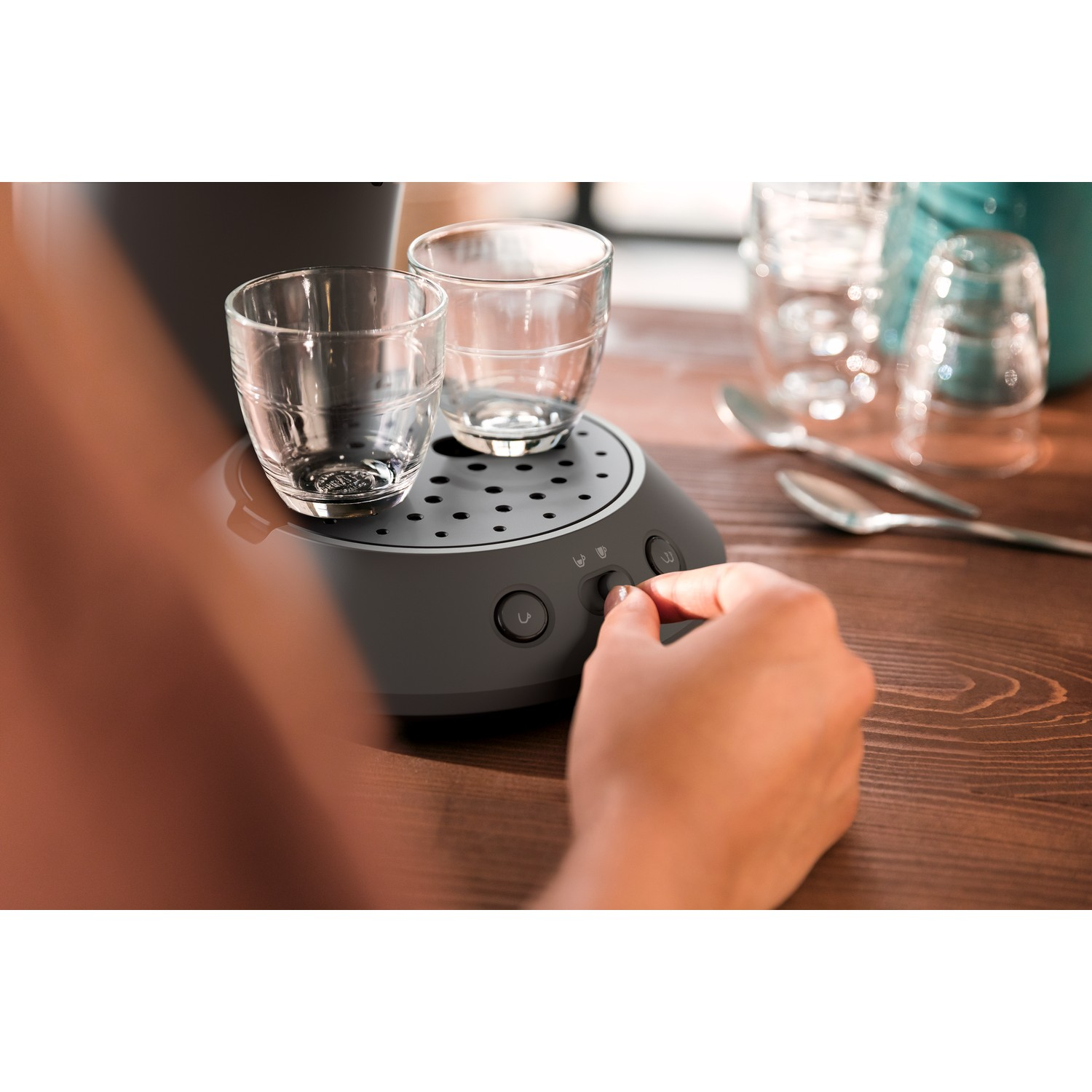 A person's hand operating a small coffee machine with two glasses on top. Other glasses and spoons are visible in the background.