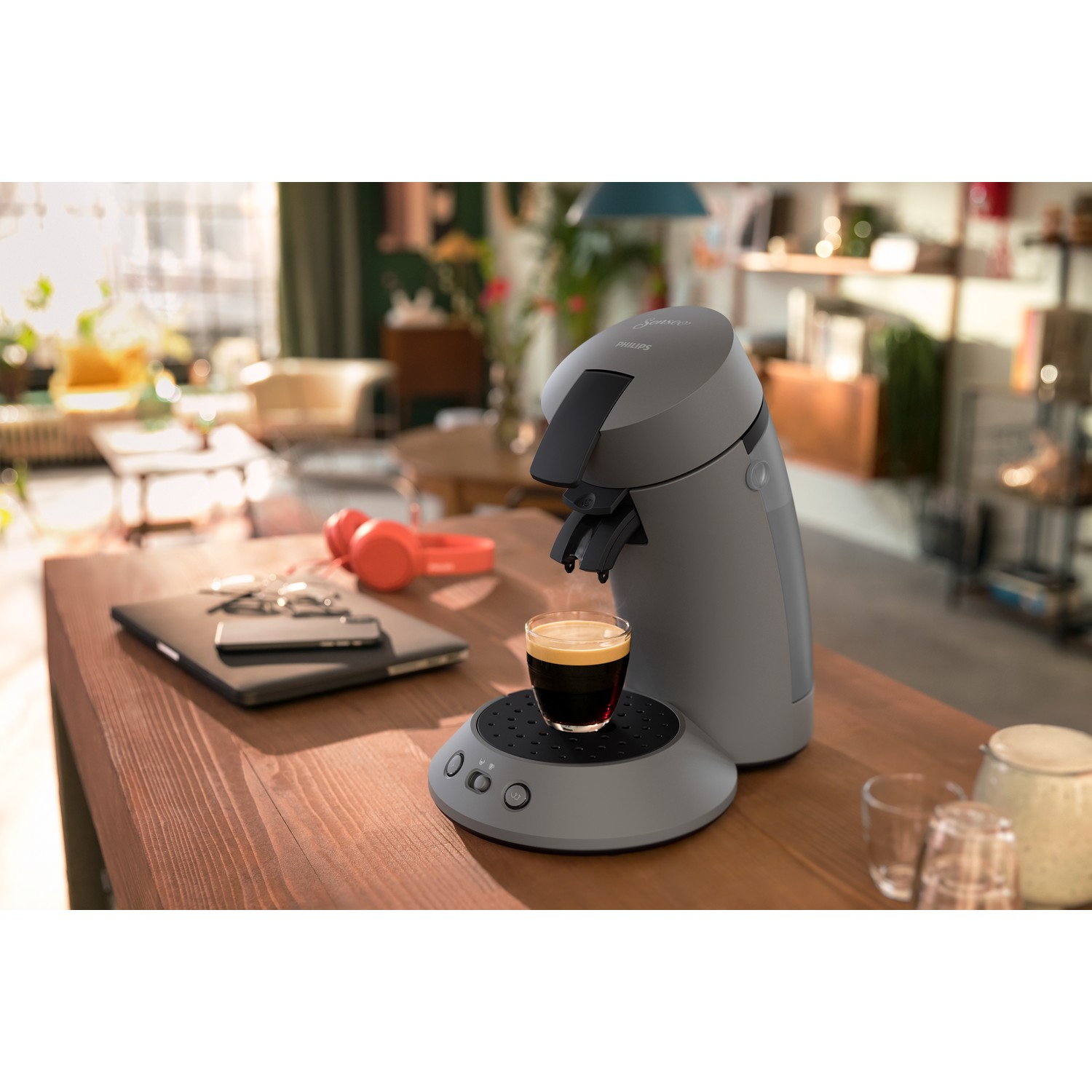 A gray Philips Senseo coffee machine brewing coffee into a glass. A laptop, phone, and headphones are on a wooden table.