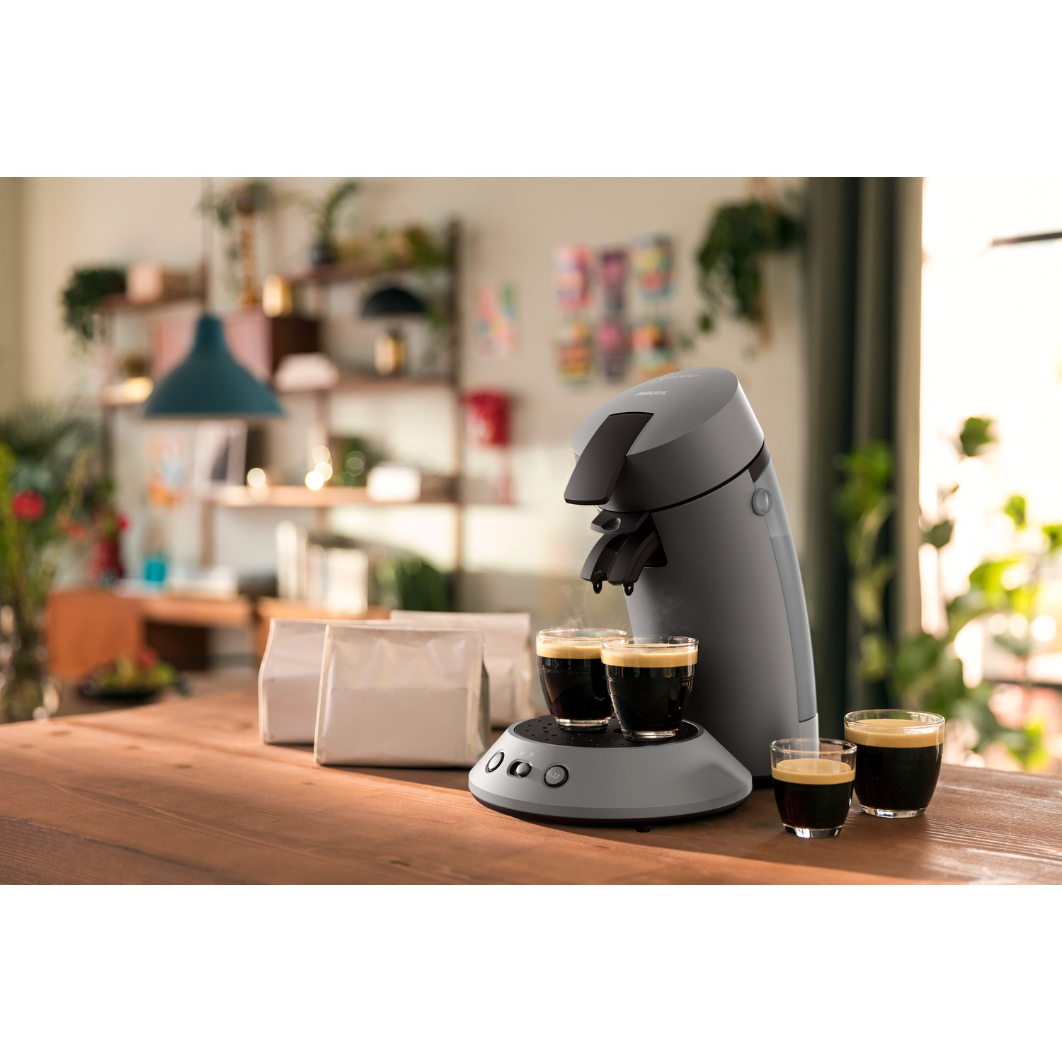 A gray coffee machine with two cups of espresso on a wooden table. Coffee bags are in the background.