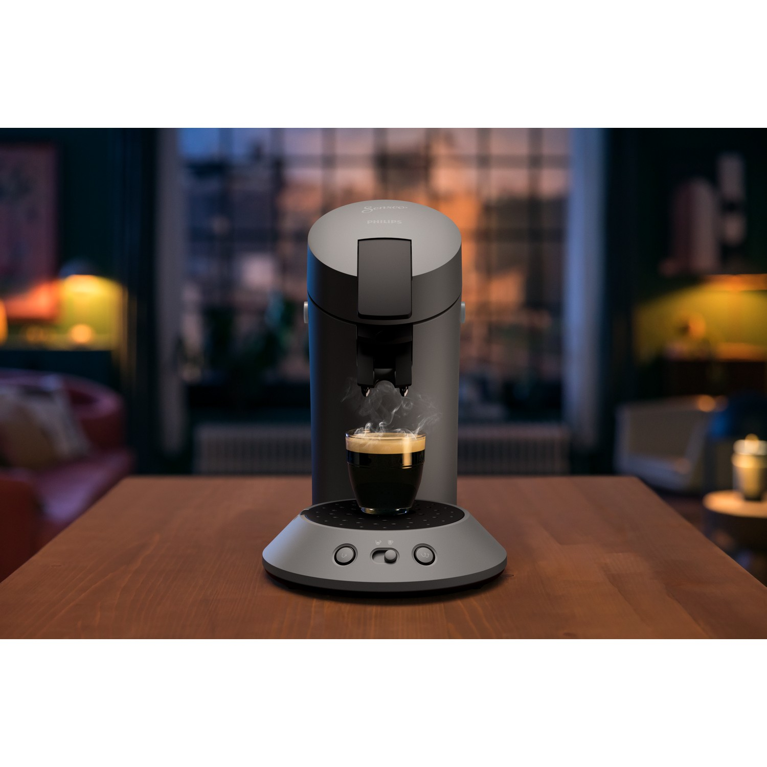 A coffee machine with a cup of coffee on a wooden table. The background shows a blurred living room.