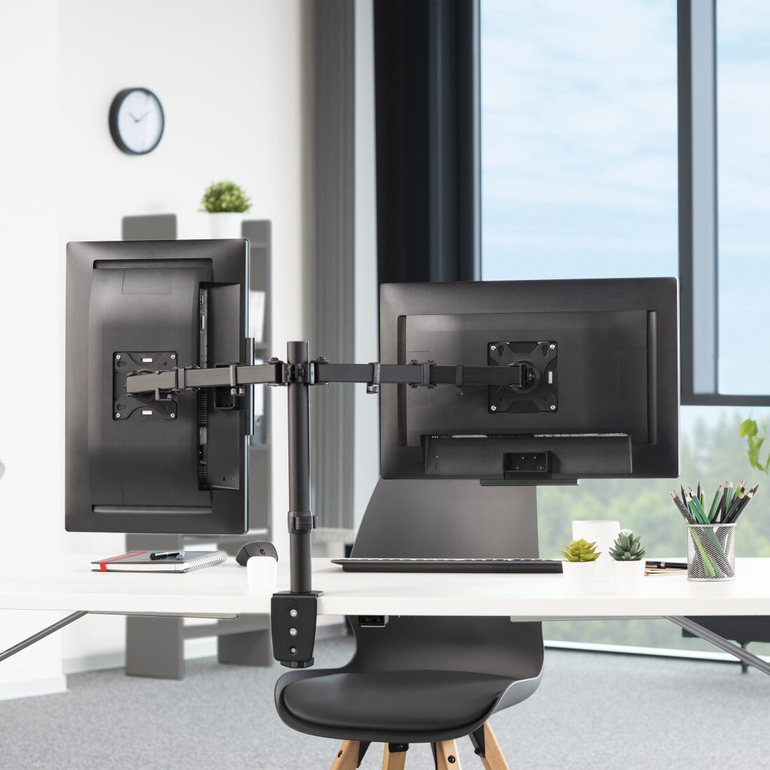 Ustawienia biurowe z dwoma monitorami na regulowanym stojaku. Widoczny biurko, zegar i rośliny.