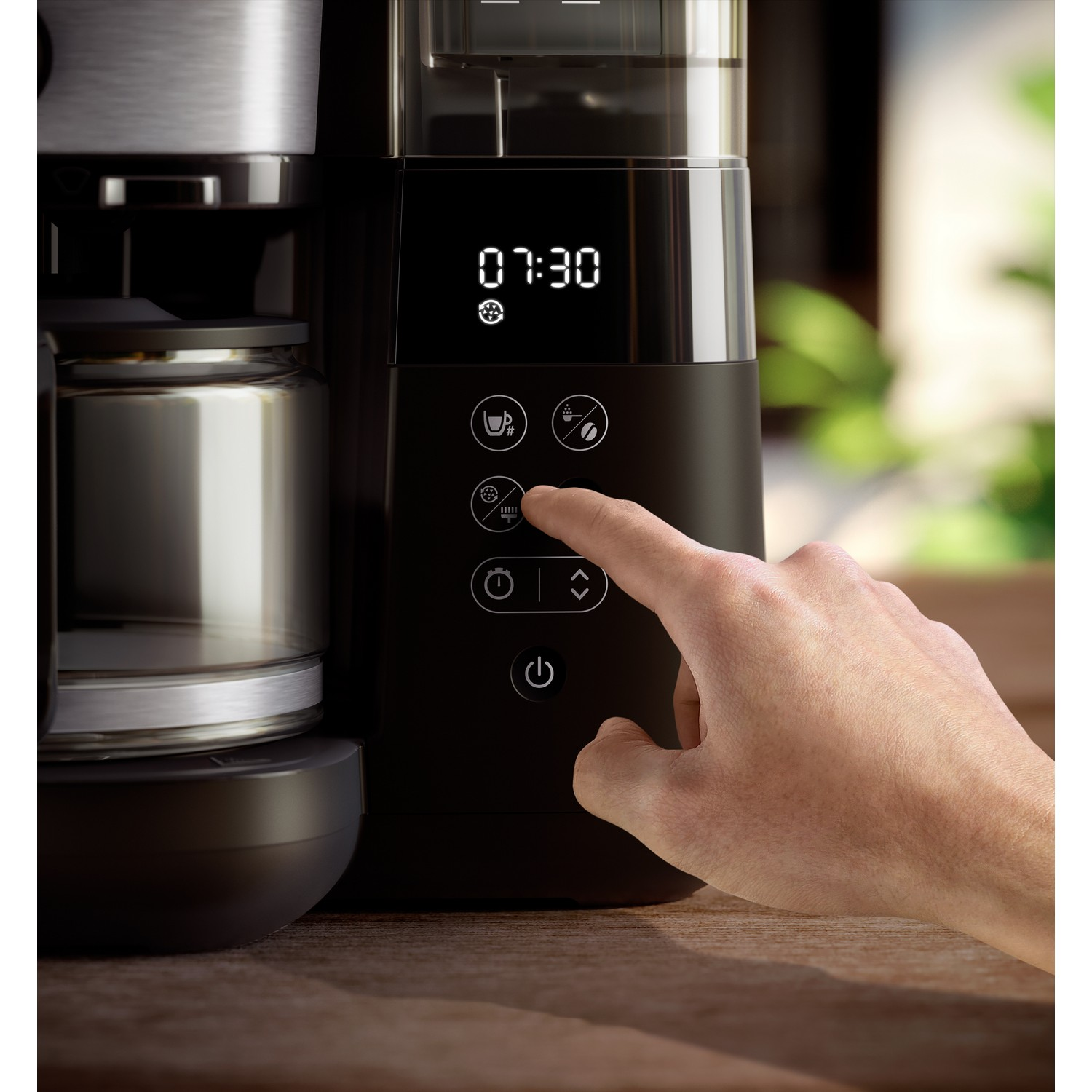 A hand pressing a button on a black coffee maker. The display shows 07:30. A glass carafe is on the left.