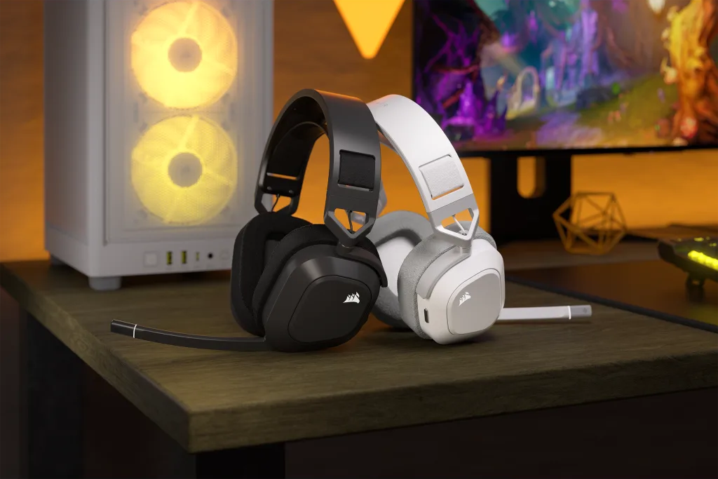 Schwarze und weiße Headsets auf einem Holztisch. Ein Computergehäuse und ein Monitor befinden sich im Hintergrund.
