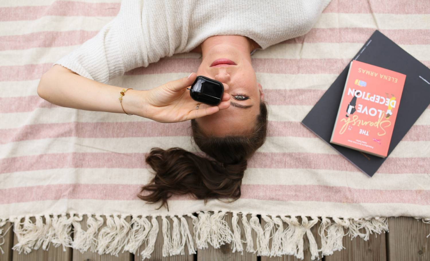Mujer acostada en manta a rayas, sosteniendo auriculares. Libro al lado.