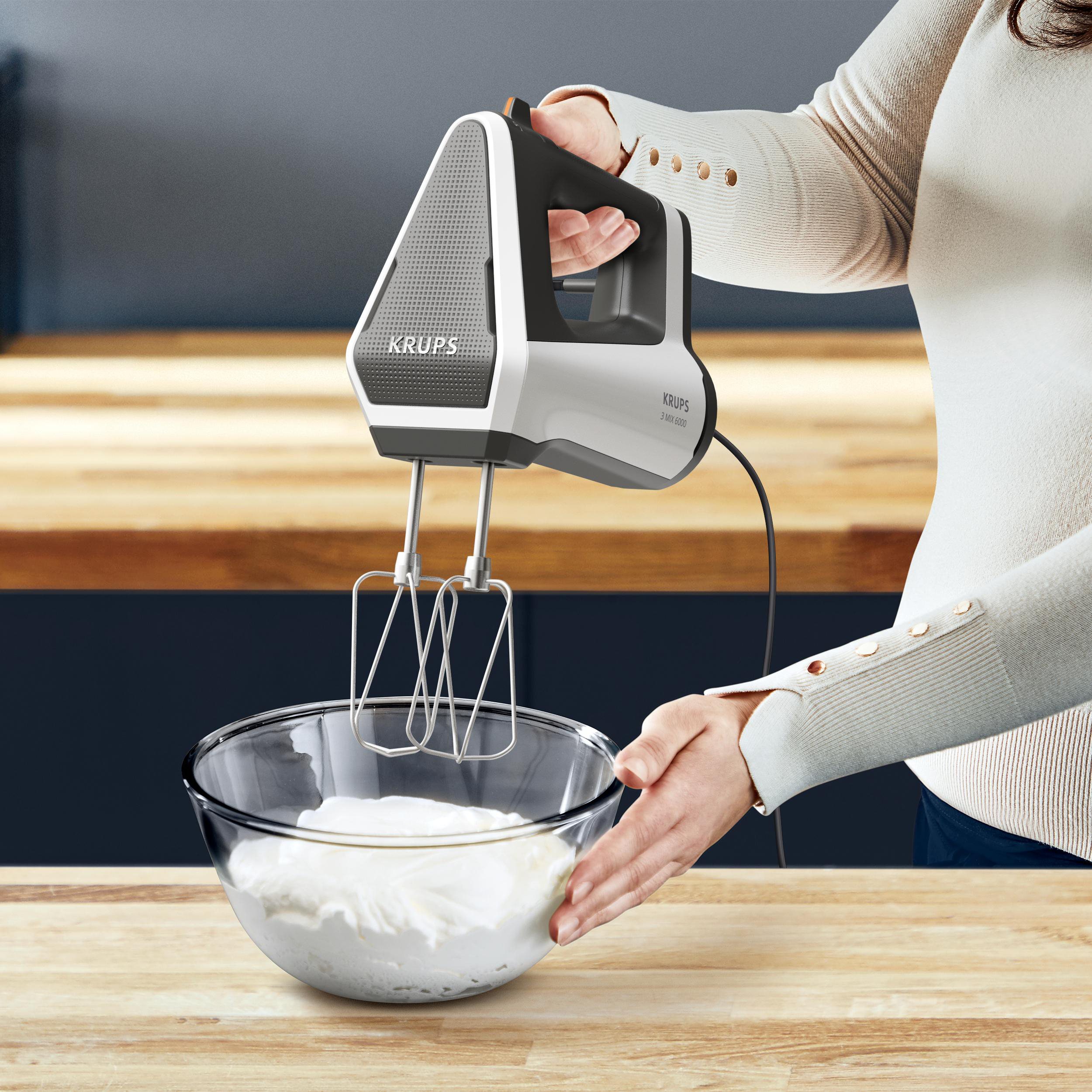 Eine Person hält einen weiß-grauen Mixer über einer Glasschüssel mit Sahne.
