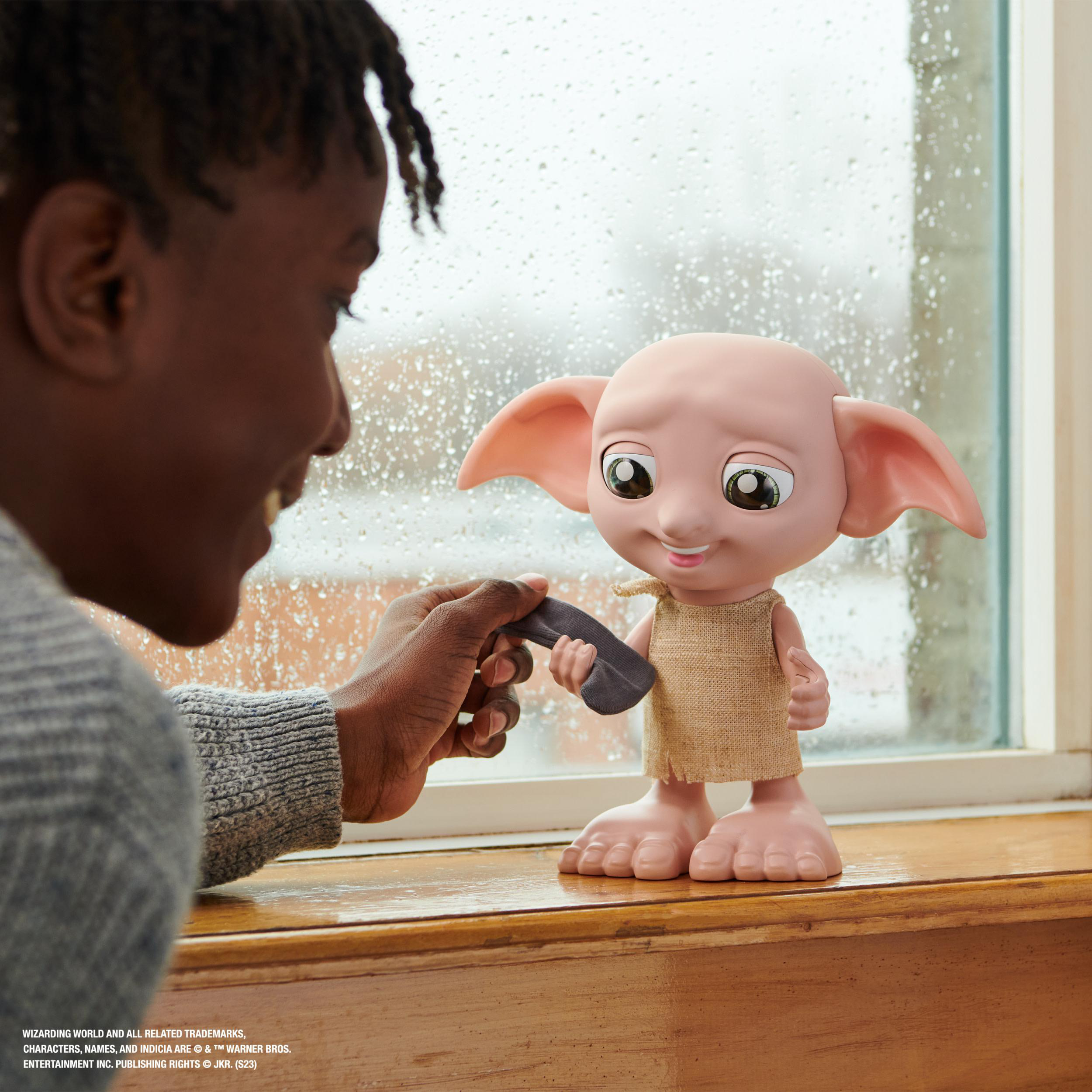 Ein Junge lächelt eine Dobby-Puppe an, die ein graues Objekt hält, in der Nähe eines Fensters mit Regentropfen.