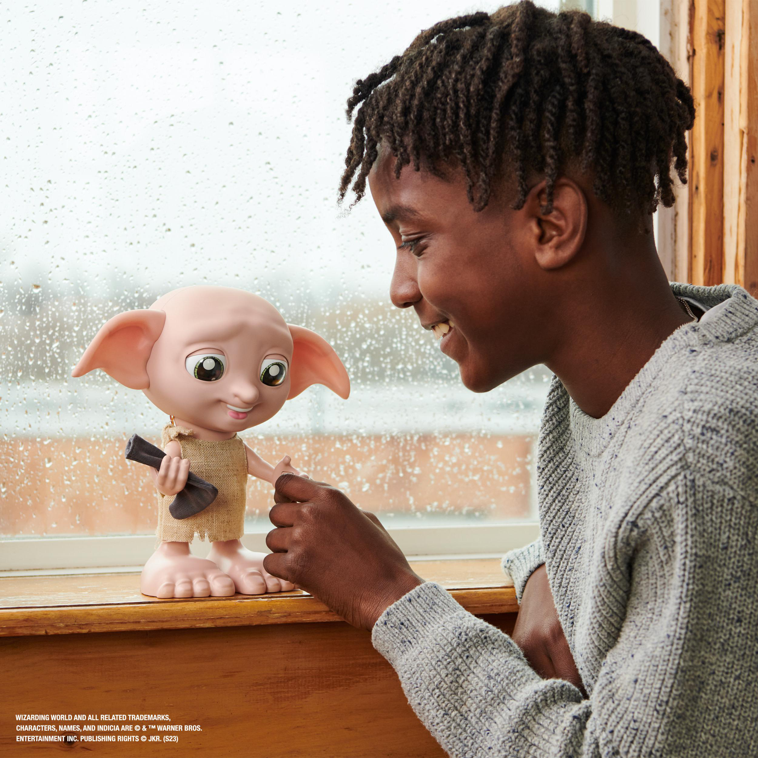 Ein Junge lächelt eine Dobby-Figur an, die eine Socke an einem regnerischen Fenster hält.