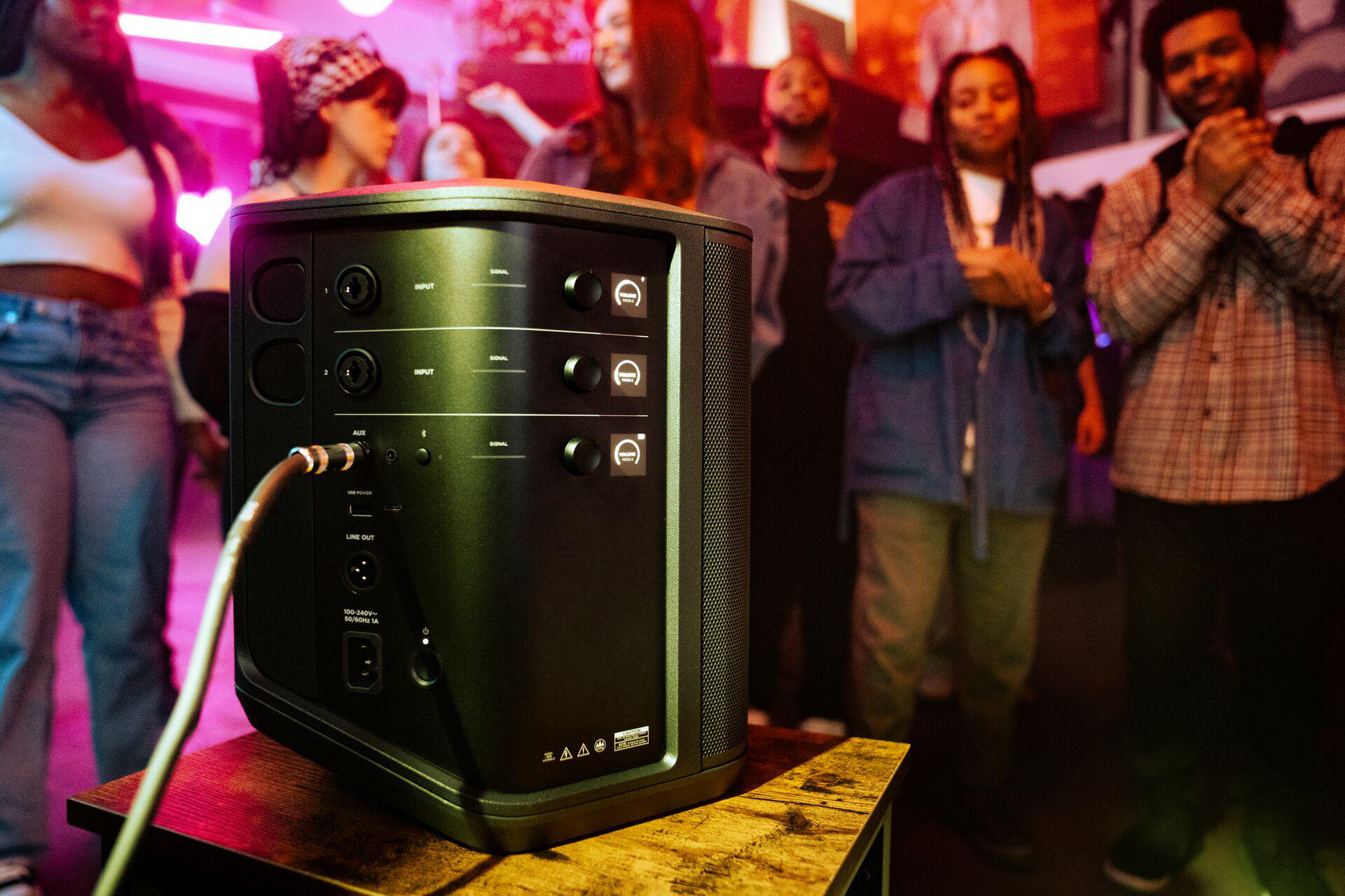 Ein schwarzer Lautsprecher auf einem Tisch mit Personen im Hintergrund. Der Lautsprecher hat mehrere Eingangsanschlüsse und Tasten.