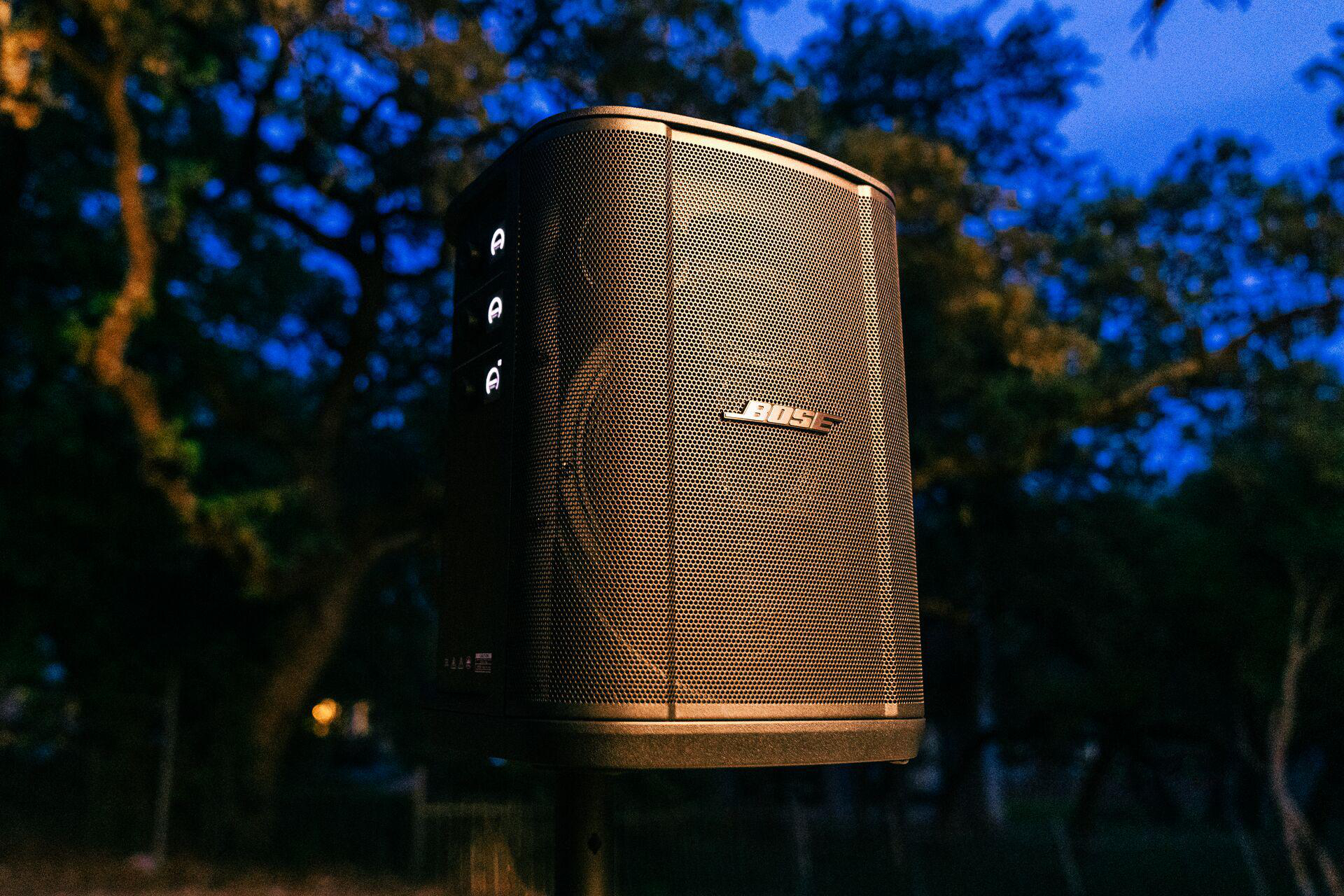 Ein Bose-Lautsprecher auf einem Ständer in einer Umgebung im Freien mit Bäumen.