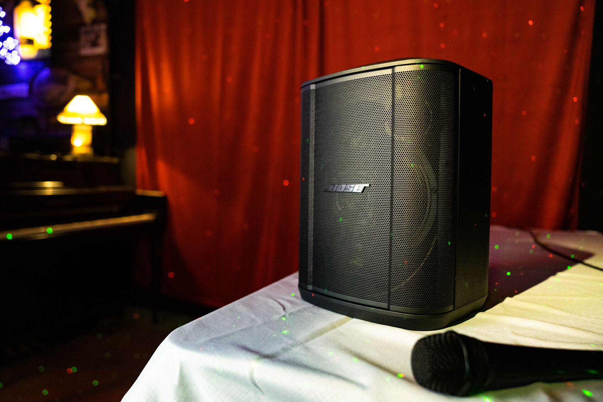 Schwarzer Bose-Lautsprecher auf einem Tisch. Mikrofon davor. Roter Vorhanghintergrund mit Lichtern.