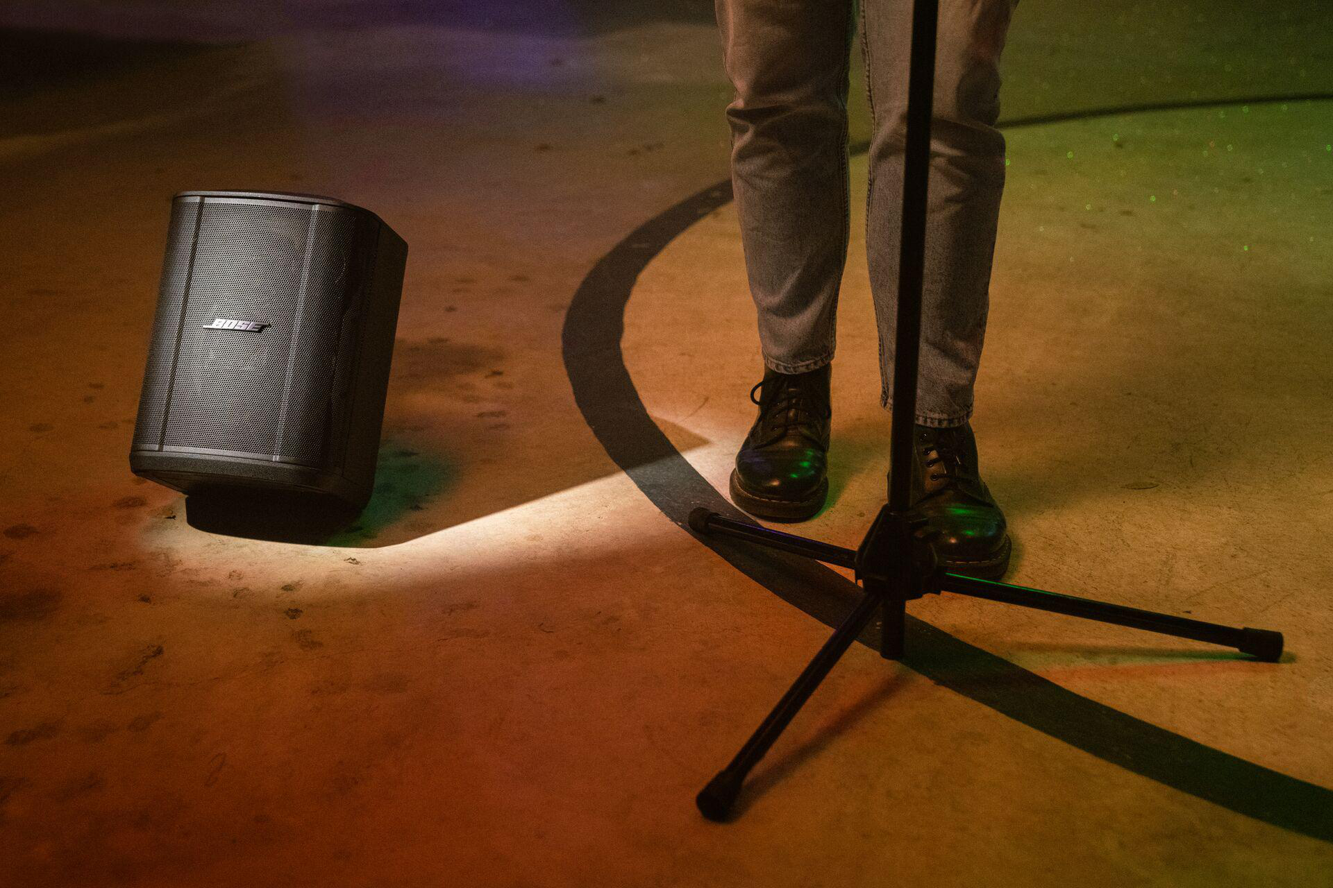 Bose-Lautsprecher neben einem Mikrofonständer. Eine Person in Jeans und Stiefeln steht daneben.