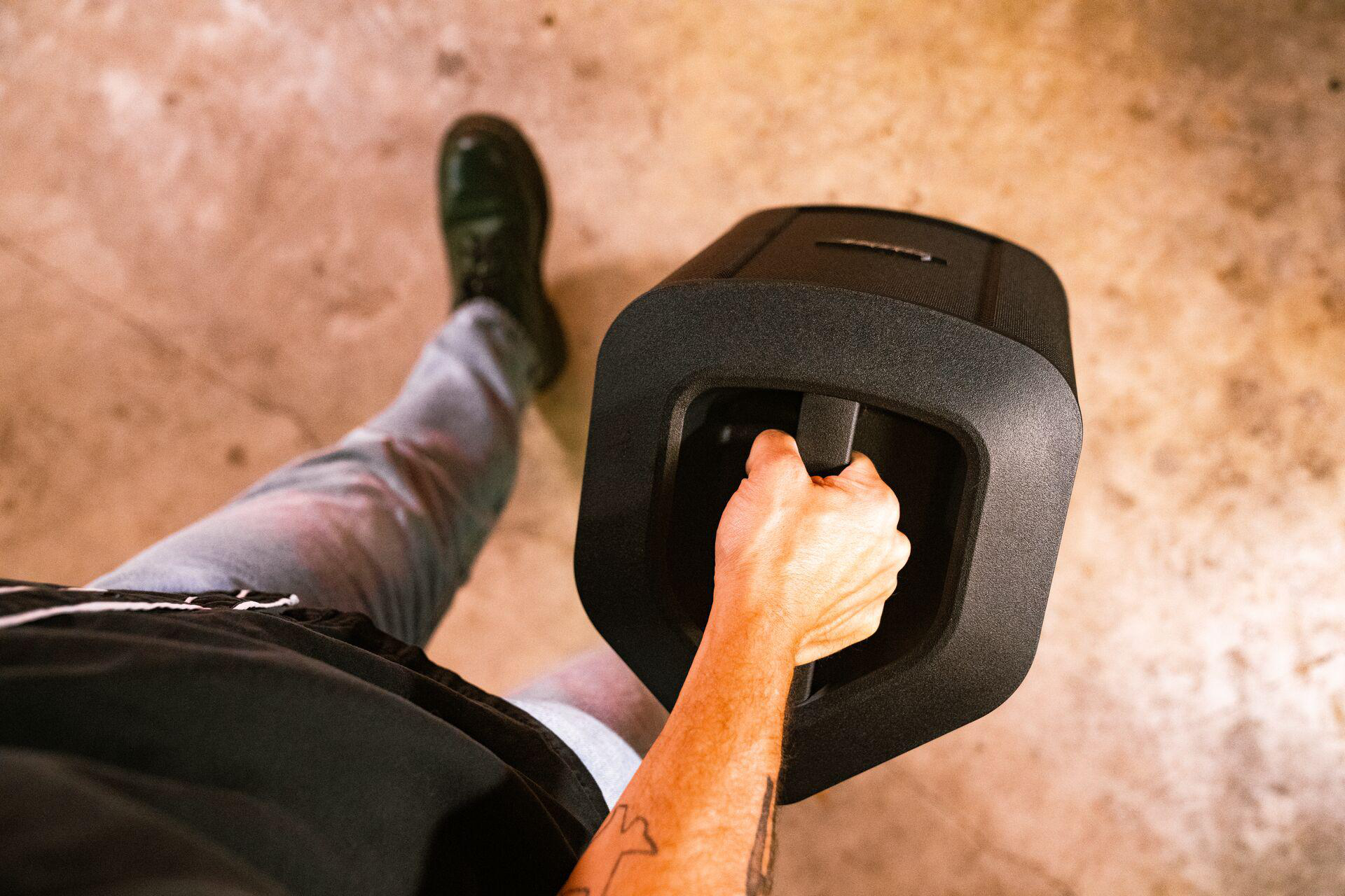 Person, die einen schwarzen Lautsprecher mit Griff hält, auf einem beige-braunen Boden.