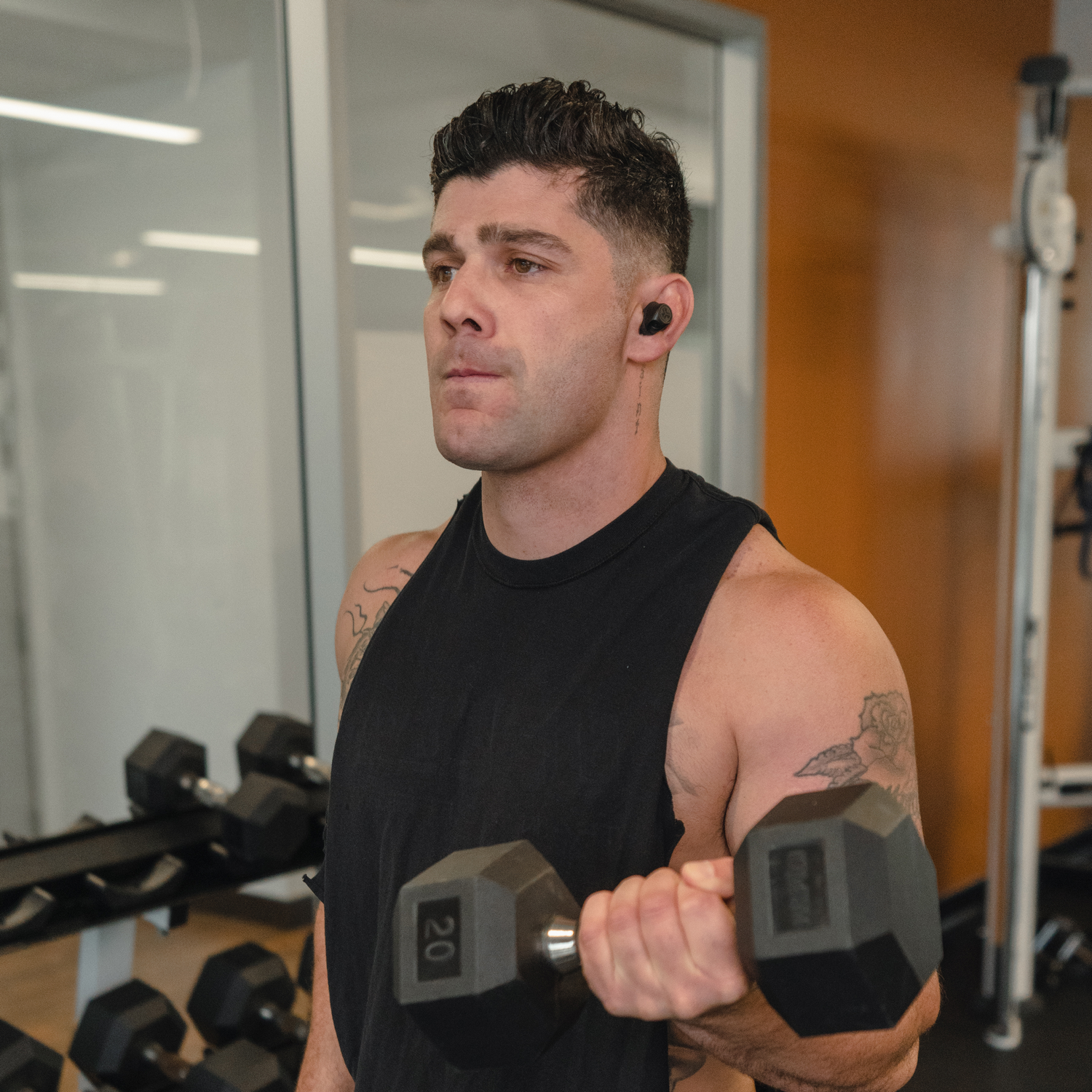 Een man is aan het trainen met een halter in een sportschool. Hij draagt een zwart tanktop en heeft tatoeages.