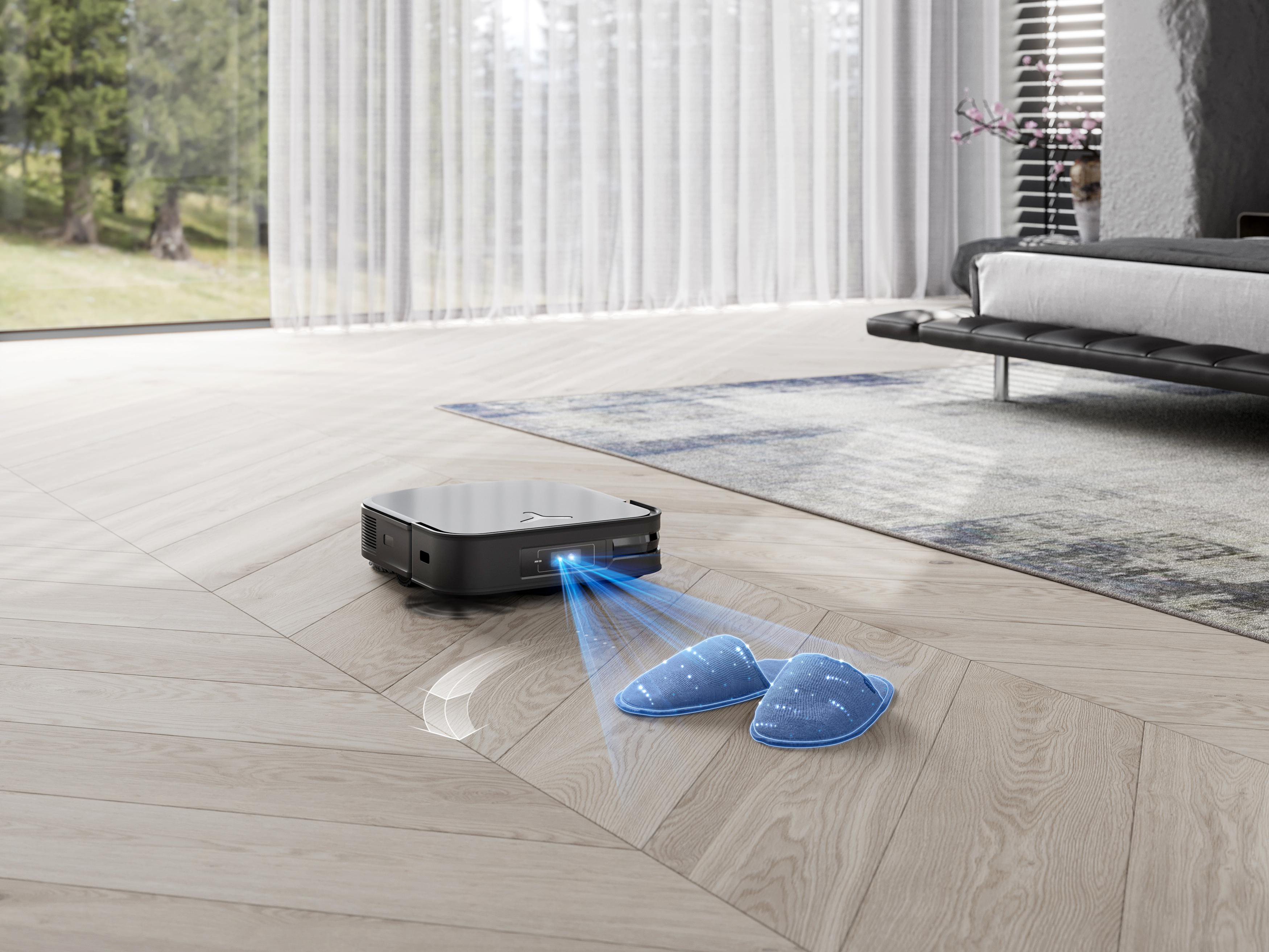 Ein Roboter-Staubsauger auf einem Holzboden, mit einem Bett im Hintergrund.