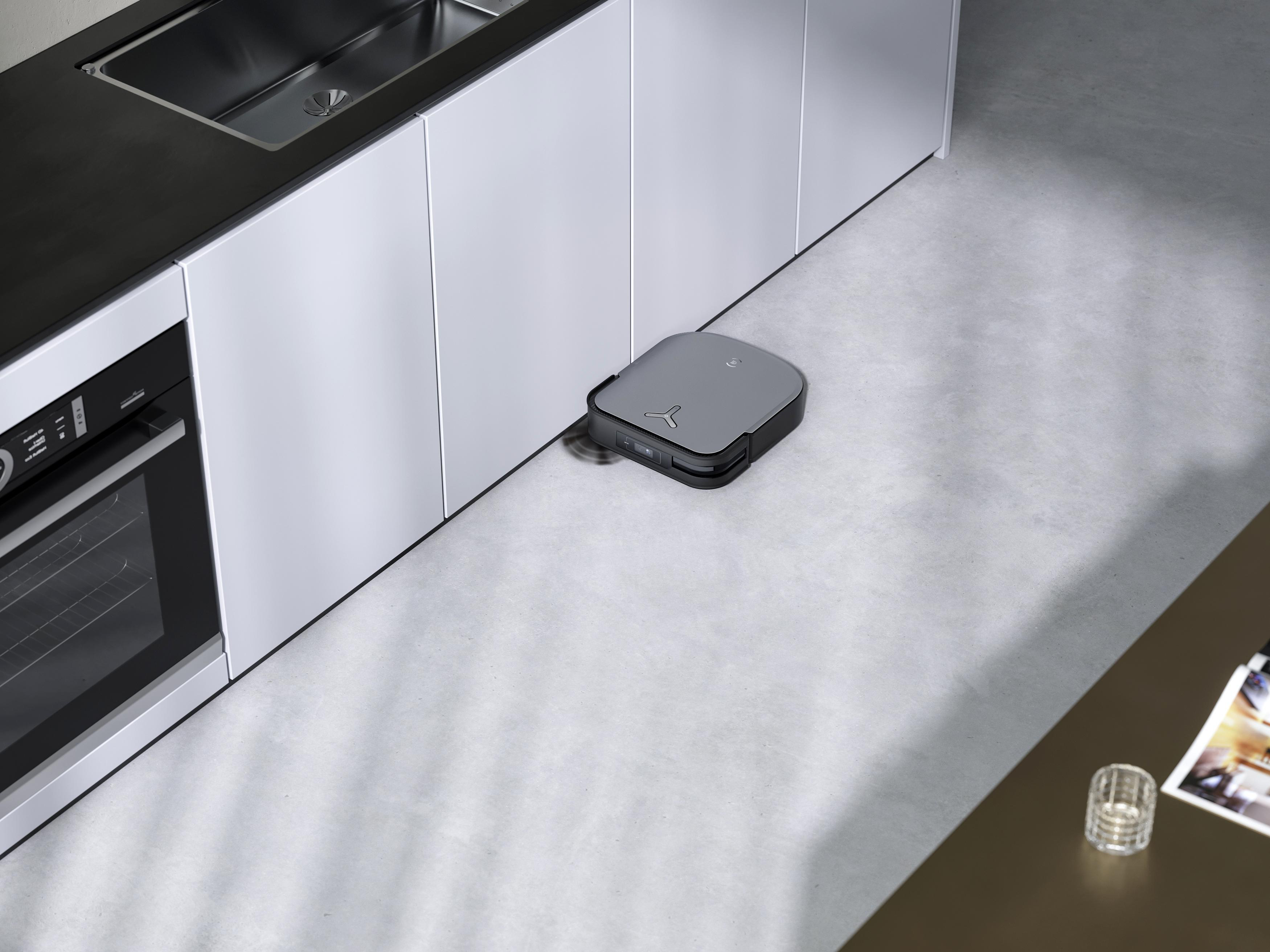 Ein Roboter-Staubsauger auf einem hellgrauen Boden in der Nähe von weißen Schränken und einer schwarzen Arbeitsplatte.