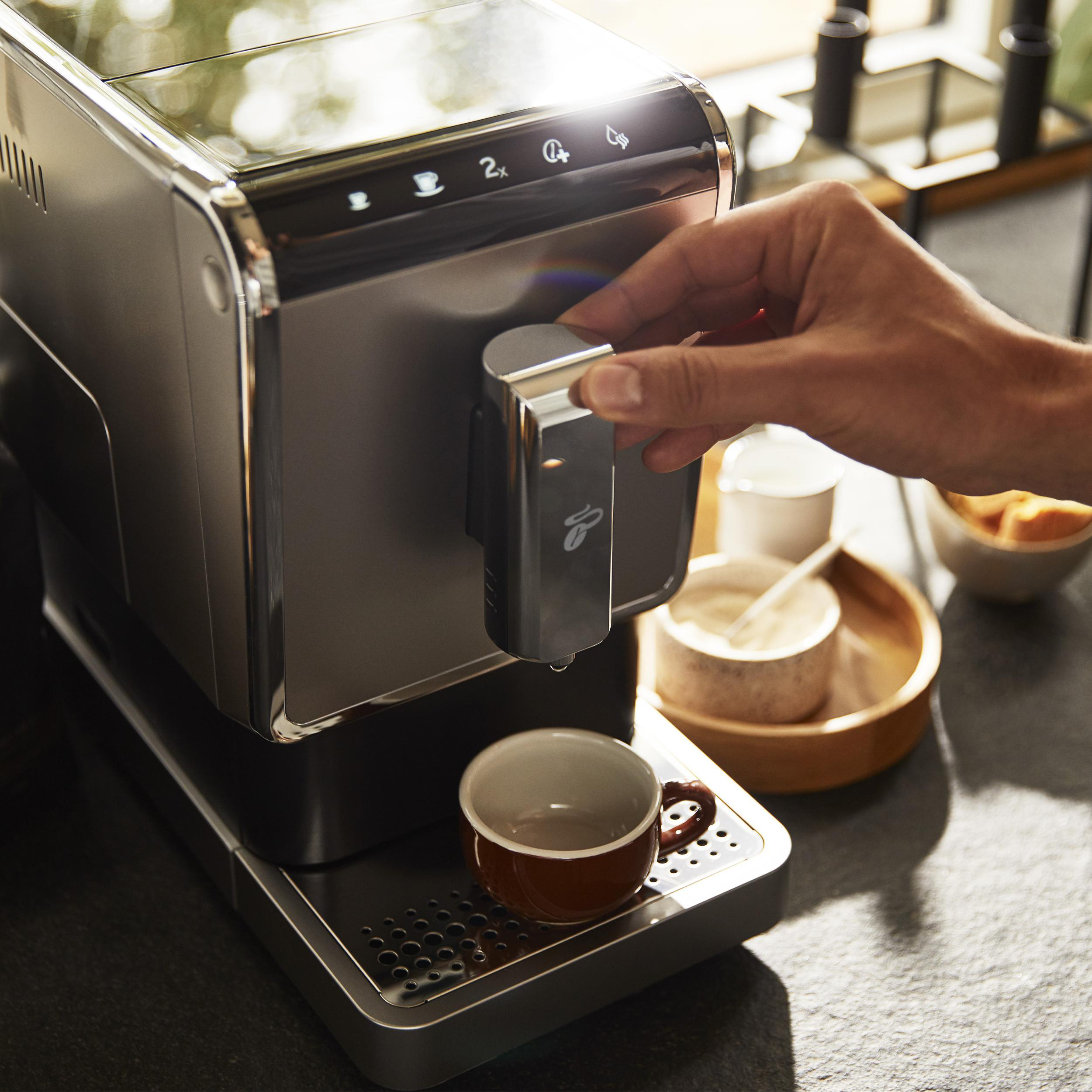 Person benutzt Kaffeemaschine. Maschine ist silber und schwarz. Tasse ist braun, Tablett schwarz.