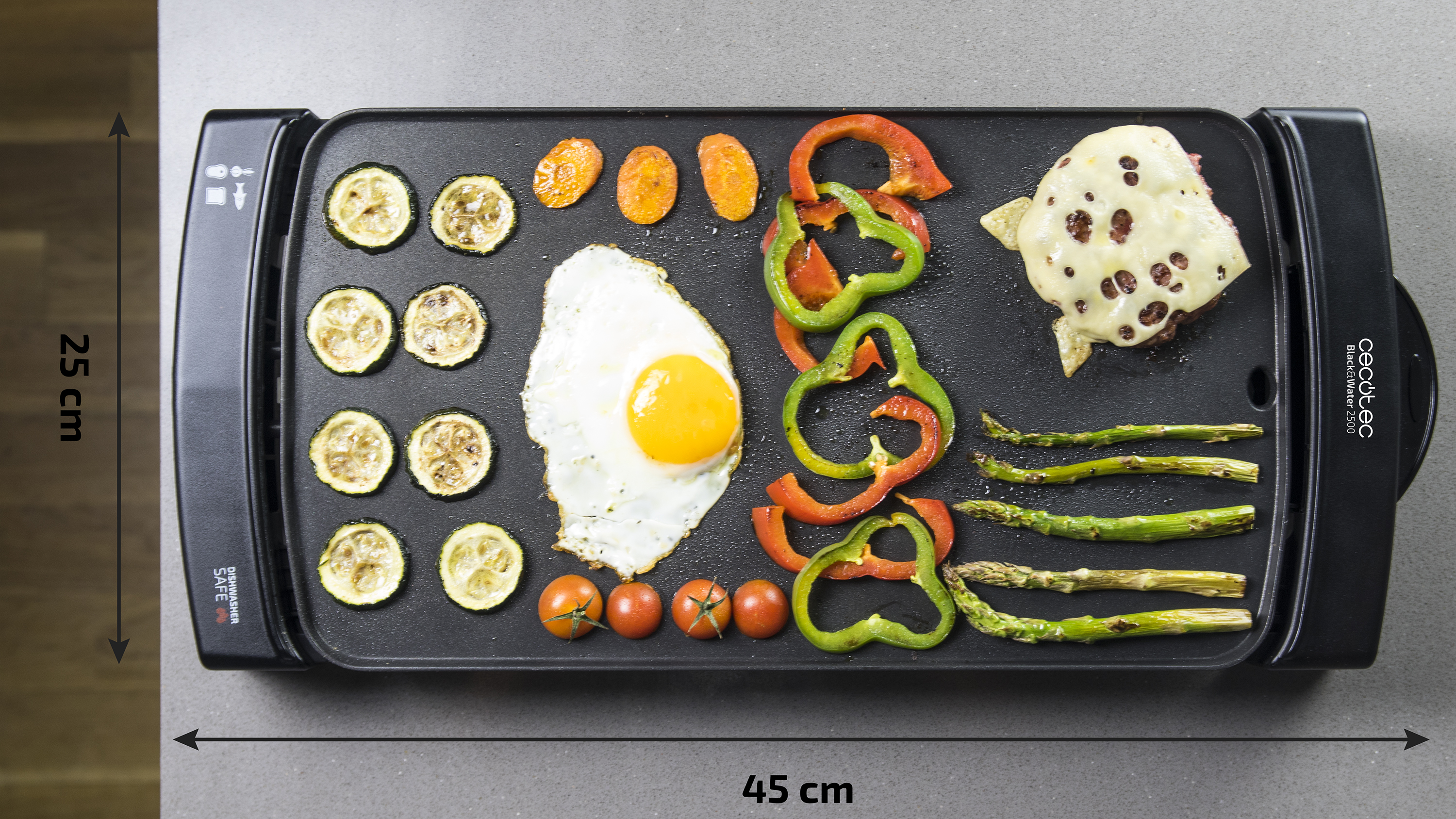 Una plancha negra con varios alimentos cocinados: calabacín, zanahorias, un huevo, tomates, pimientos, una hamburguesa y espárragos.