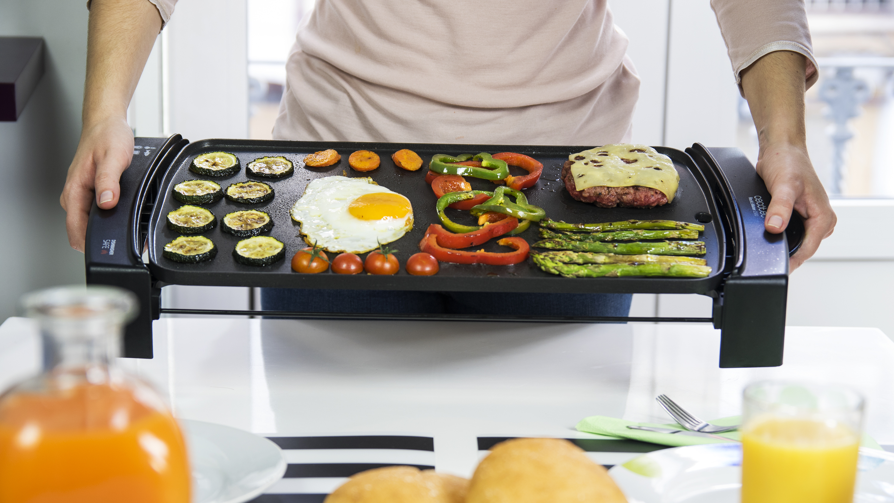 Una persona sostiene una plancha con comida cocinada: calabacín, zanahorias, pimientos, huevo, hamburguesa, espárragos y tomates.